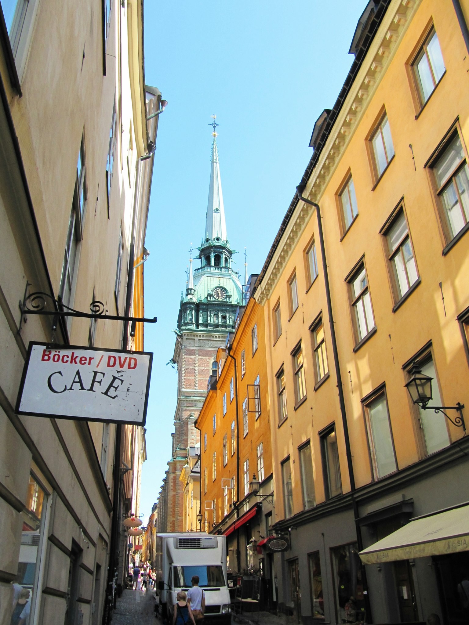 Narrow street with a cafe sign and yellow buildings featuring a tower with a spire in the background