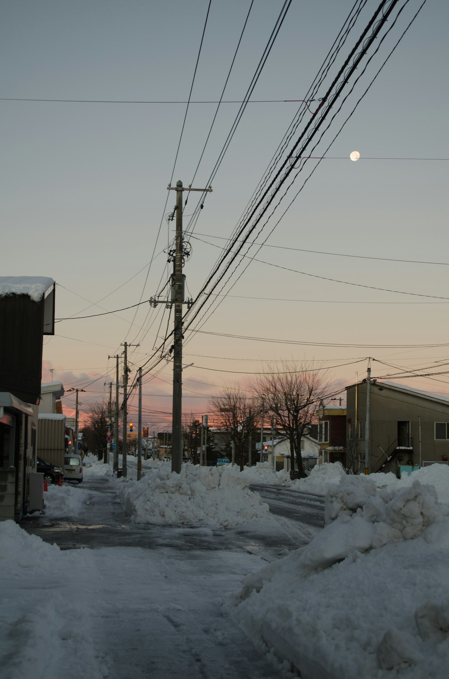 Calle cubierta de nieve con postes de electricidad y cielo al atardecer con la luna