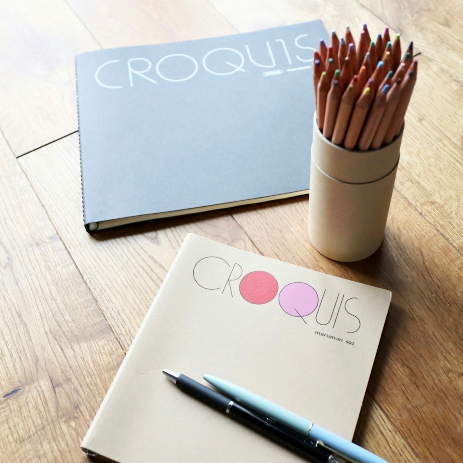 Two sketchbooks and a container of colored pencils on a wooden floor
