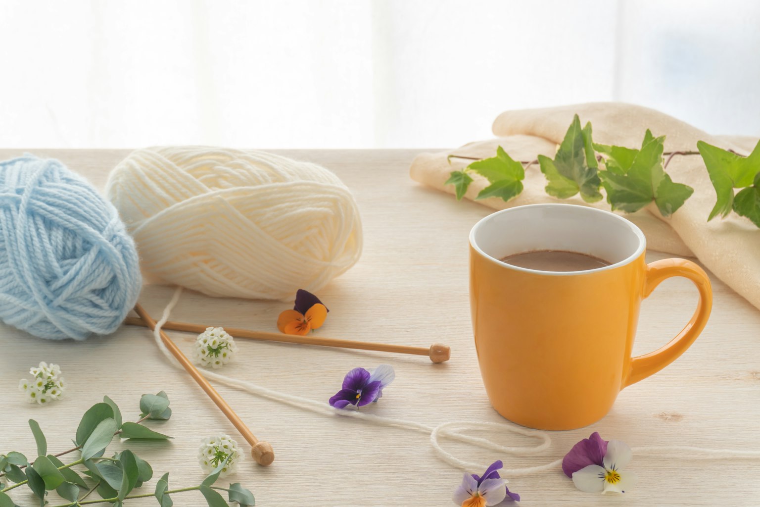 Una escena interior brillante con una taza amarilla, ovillos de lana y flores y hojas esparcidas