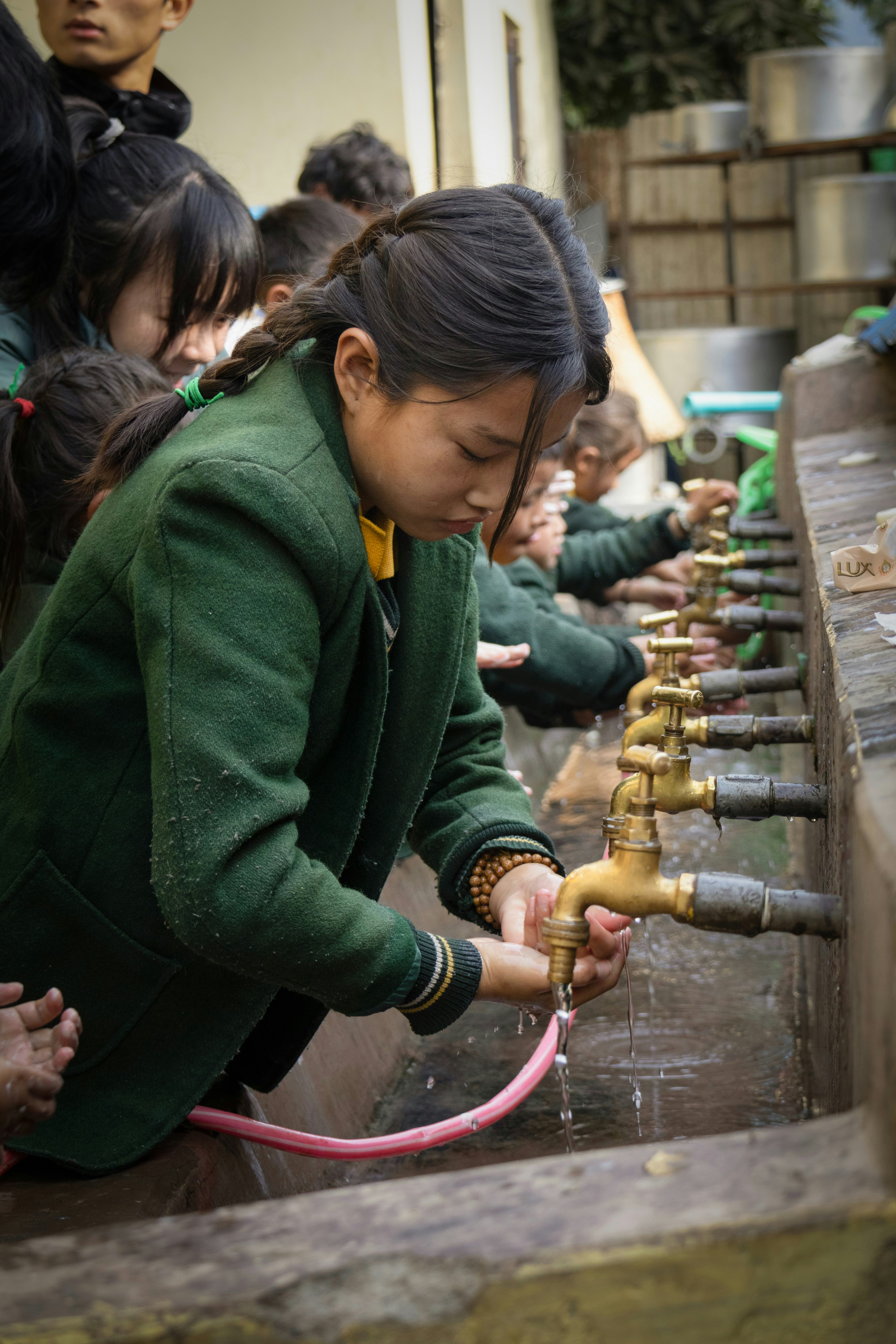 路地の屋外で並ぶ子どもたちが水道の蛇口を回す体験風景