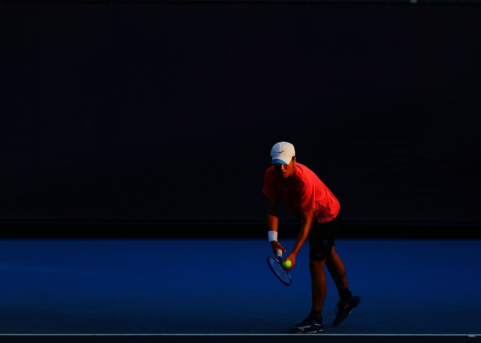 Tennis Spieler bereitet sich auf den Aufschlag vor trägt ein rotes Shirt und eine weiße Kappe blauer Platz und dunkler Hintergrund