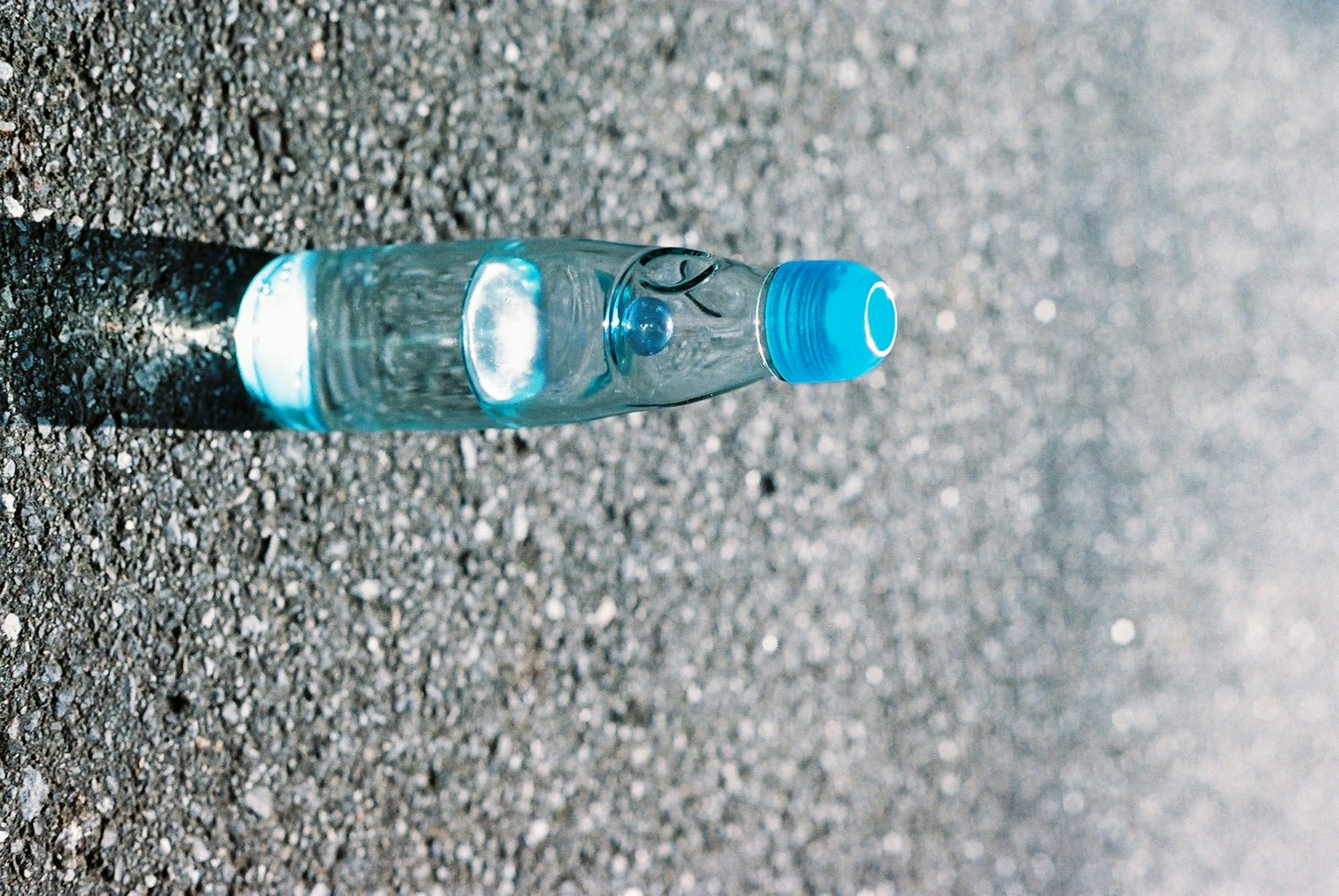Eine transparente Wasserflasche mit blauem Deckel liegt auf Asphalt
