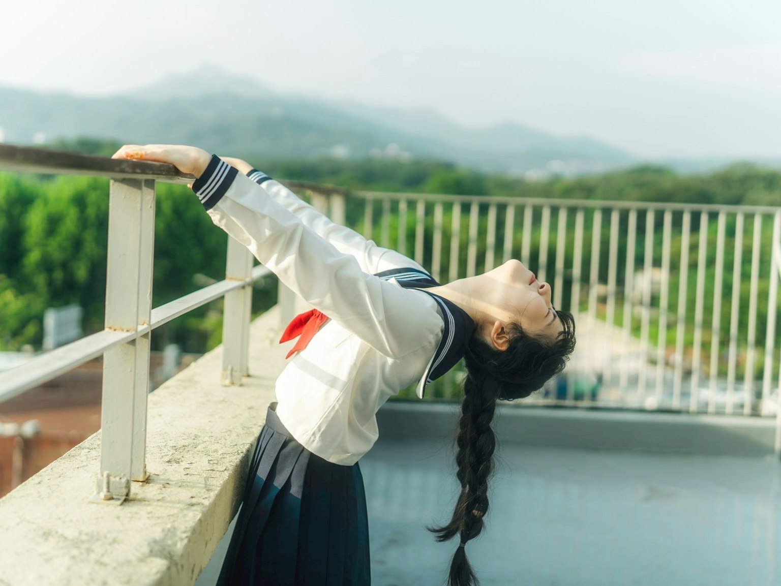 屋上で背を反らせている女子学生の姿 緑の風景と青空が背景