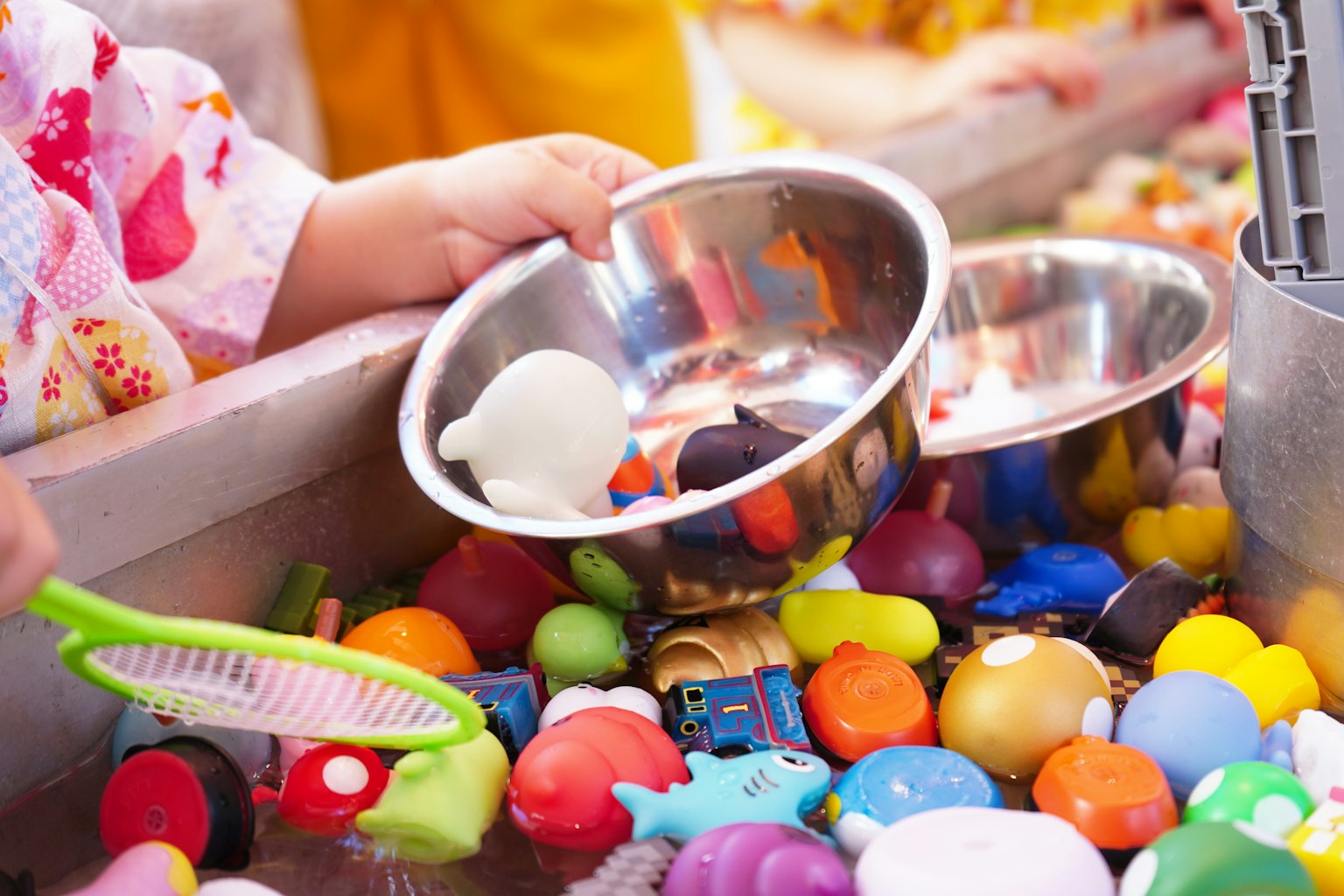 A child playing in a water-filled area with colorful toys using a silver bowl to scoop some toys