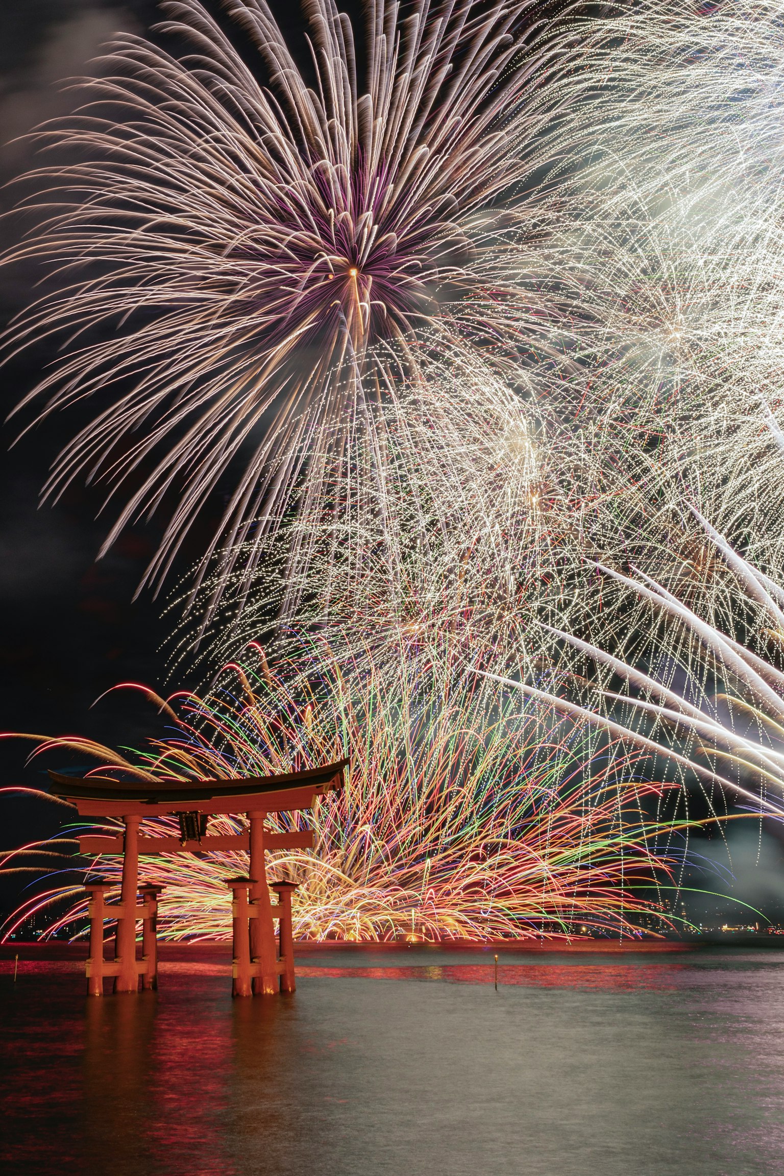 夜空に花火が打ち上げられ、赤い鳥居が水面に映る美しい風景