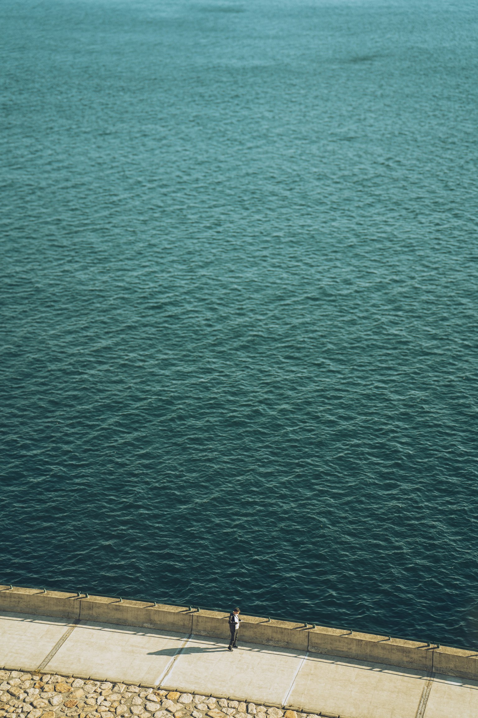 Eine einsame Figur steht auf einem Gehweg am Meer mit einer blauen Wasseroberfläche