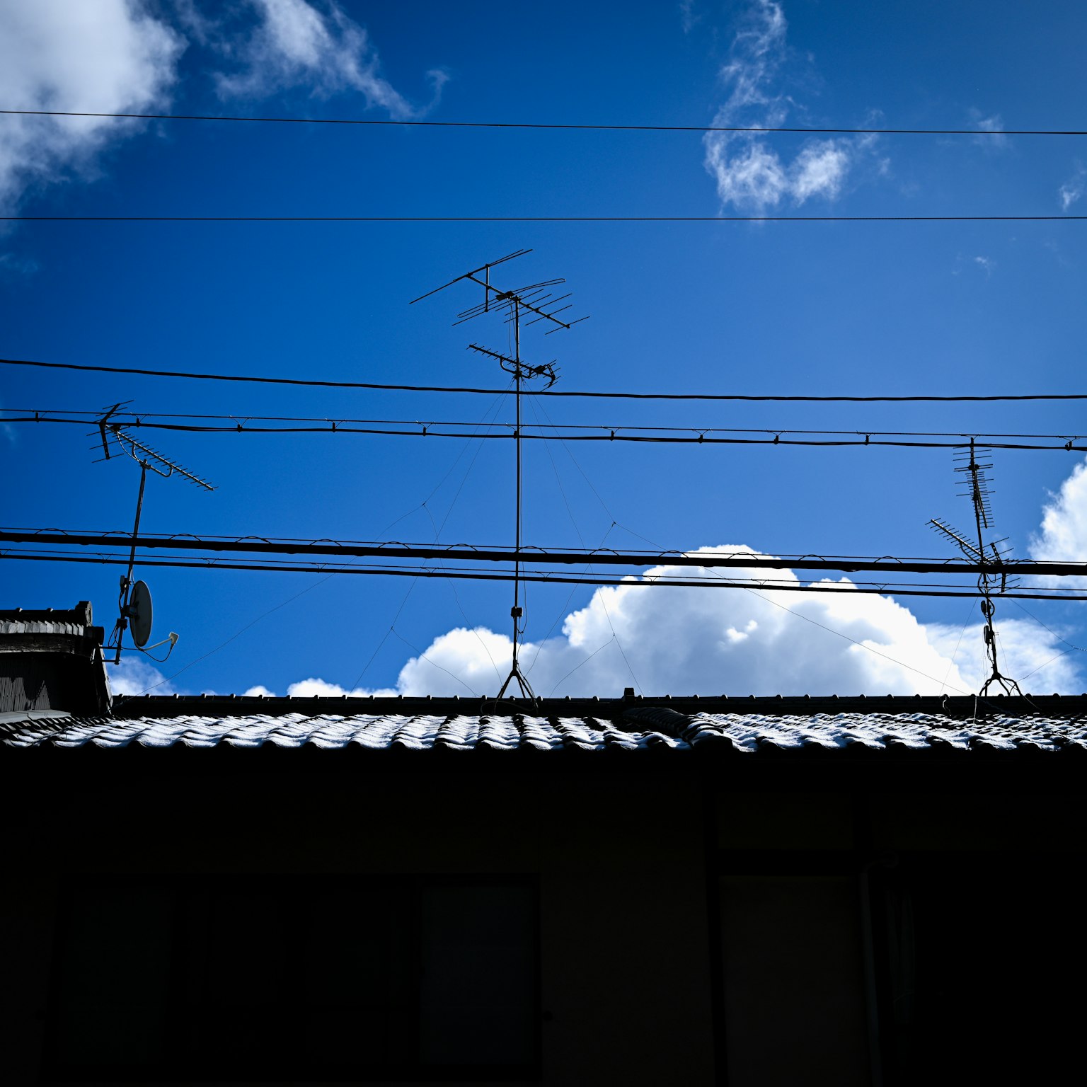A view of a house roof and electrical wires against a blue sky with white clouds