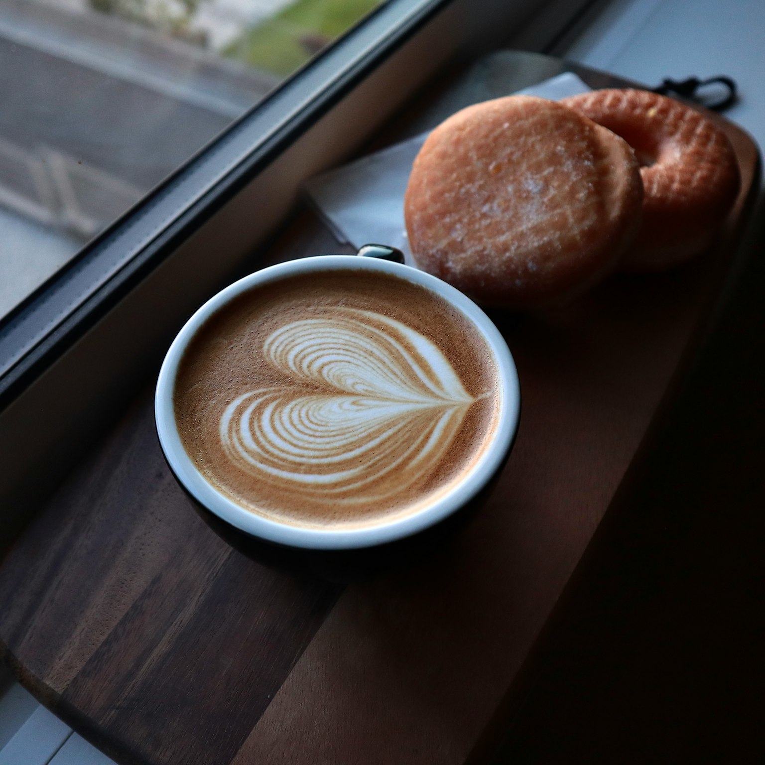 Latte dengan seni latte berbentuk hati di samping kue di papan kayu dekat jendela