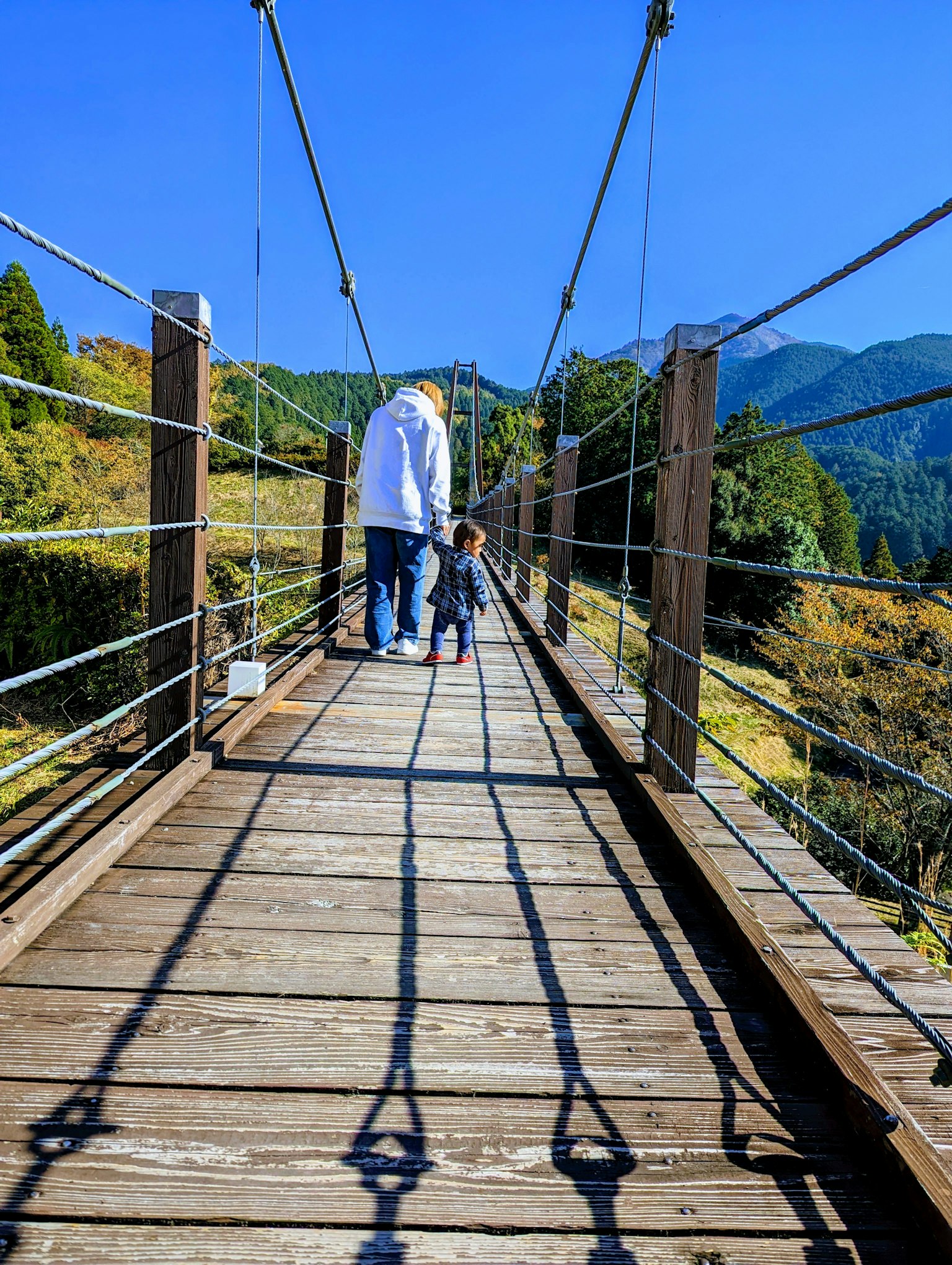 Adulto e bambino che camminano su un ponte sospeso con montagne e cielo blu sullo sfondo ombre suggestive