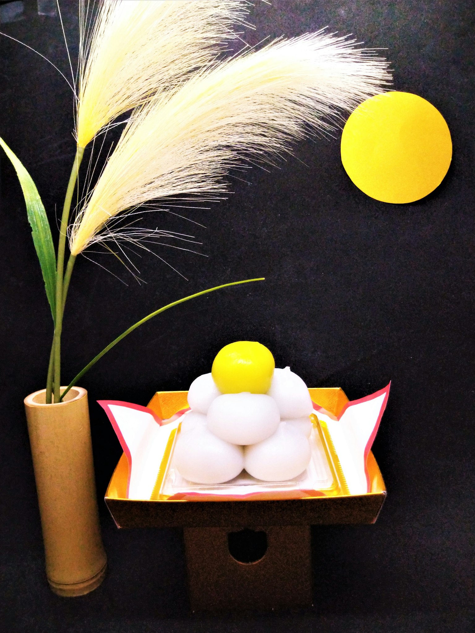 A decorative display of stacked white rice cakes with a yellow rice cake on top and pampas grass in a vase