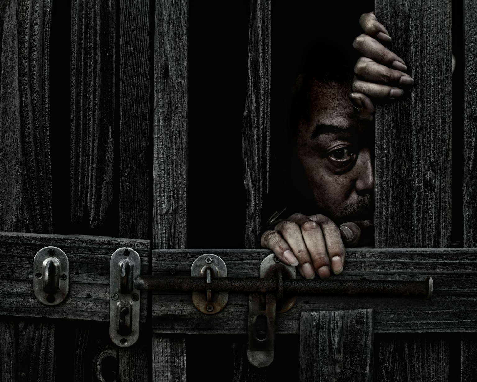 A person's eye and hand visible through a gap in a wooden door creating a sense of tension