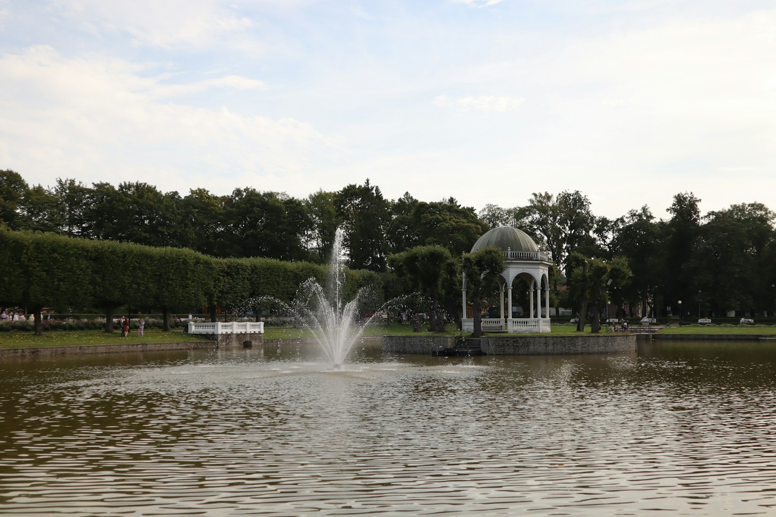 Una fuente en el centro de un estanque rodeada de vegetación exuberante y un gazebo blanco al fondo