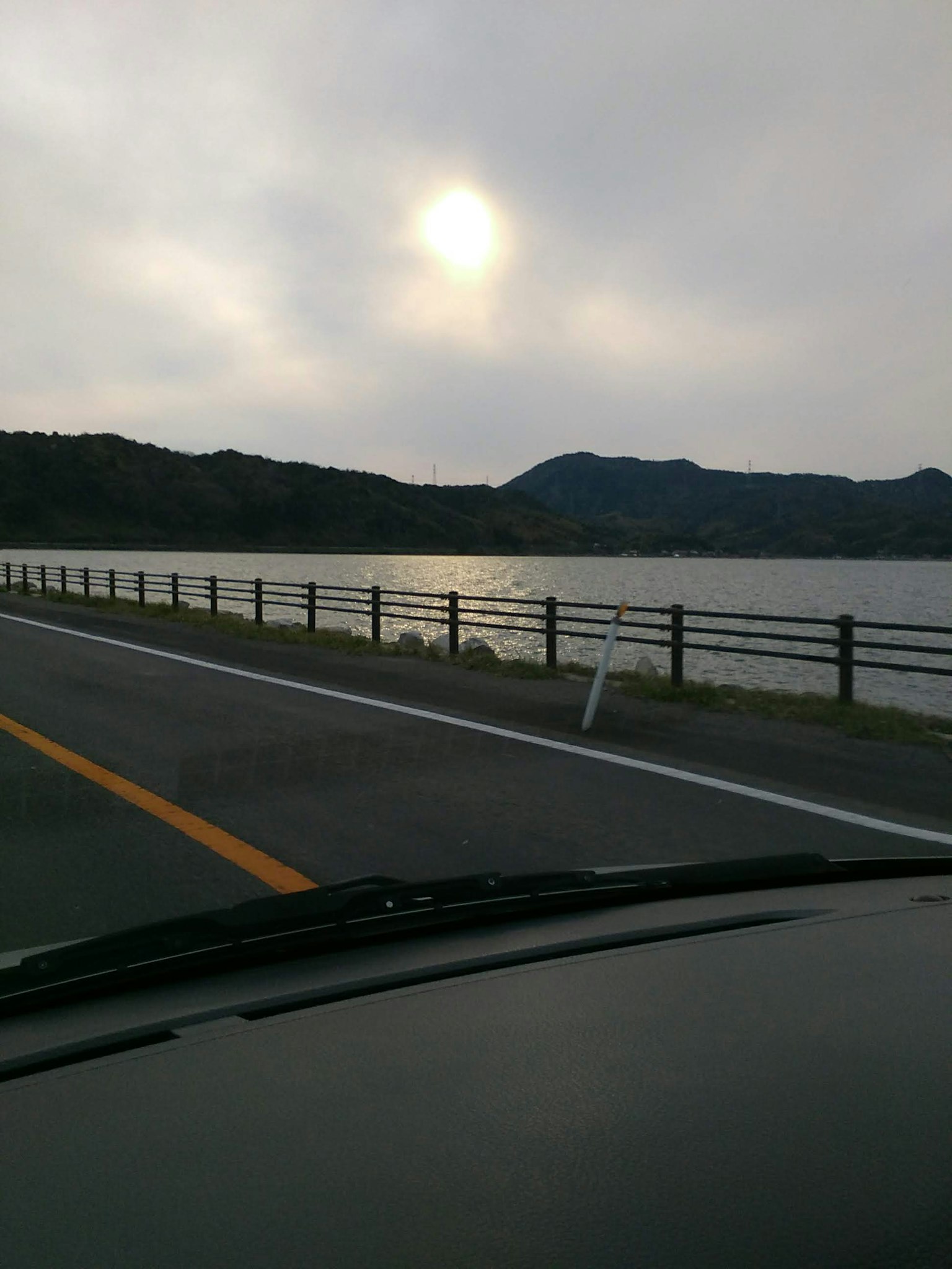 Vista panoramica di un lago e montagne sotto un cielo nuvoloso con una recinzione lungo la strada e il cruscotto di un'auto visibile