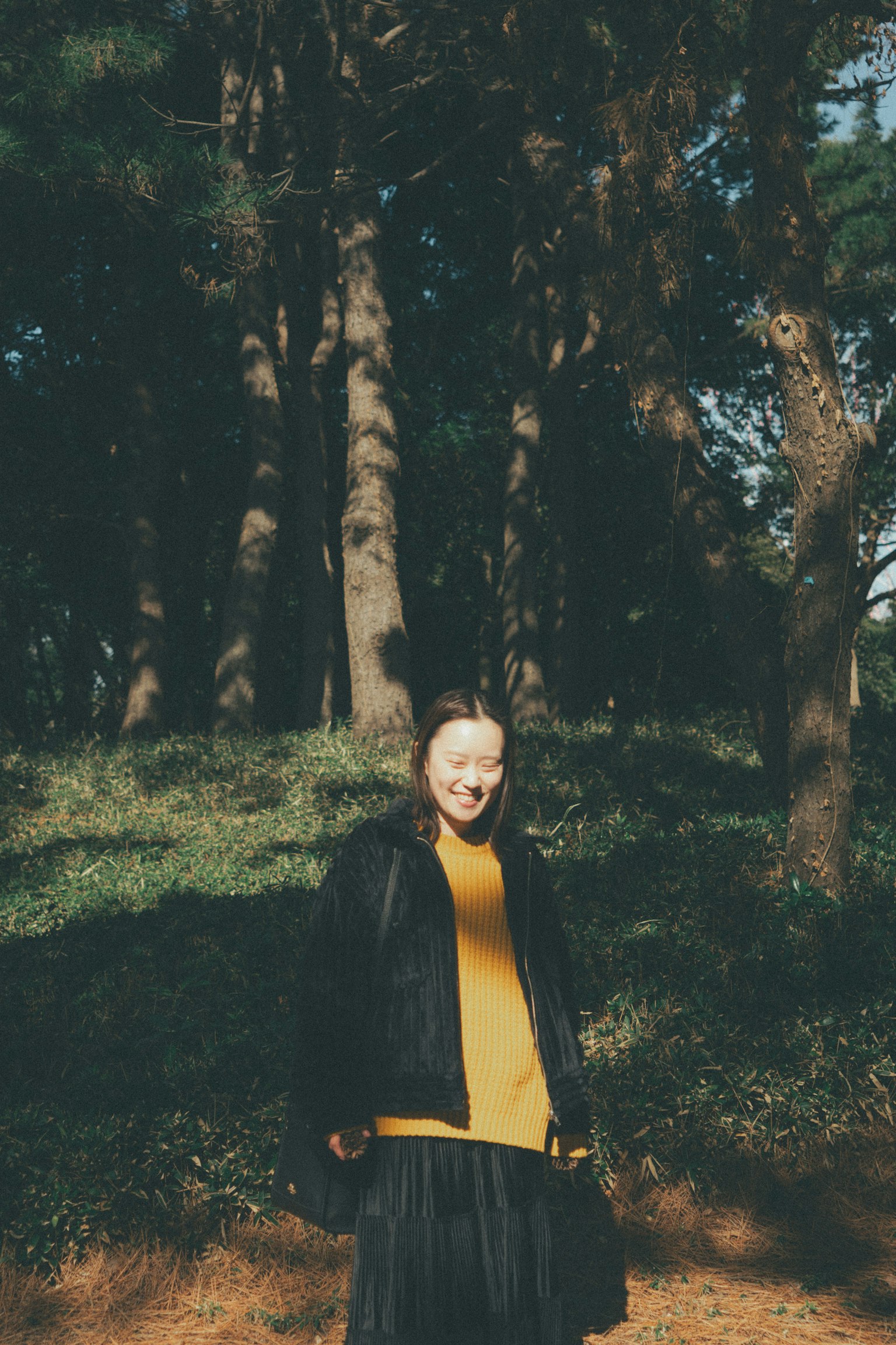 Foto wanita tersenyum di hutan mengenakan sweter kuning cerah dan jaket hitam