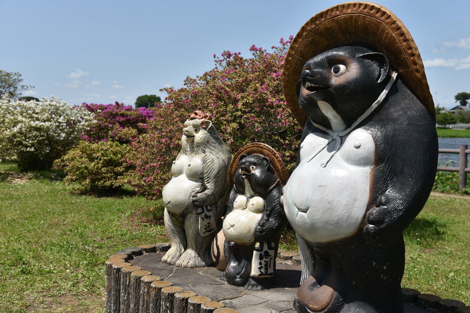 Tres estatuas de tanuki de pie en un jardín con flores coloridas al fondo
