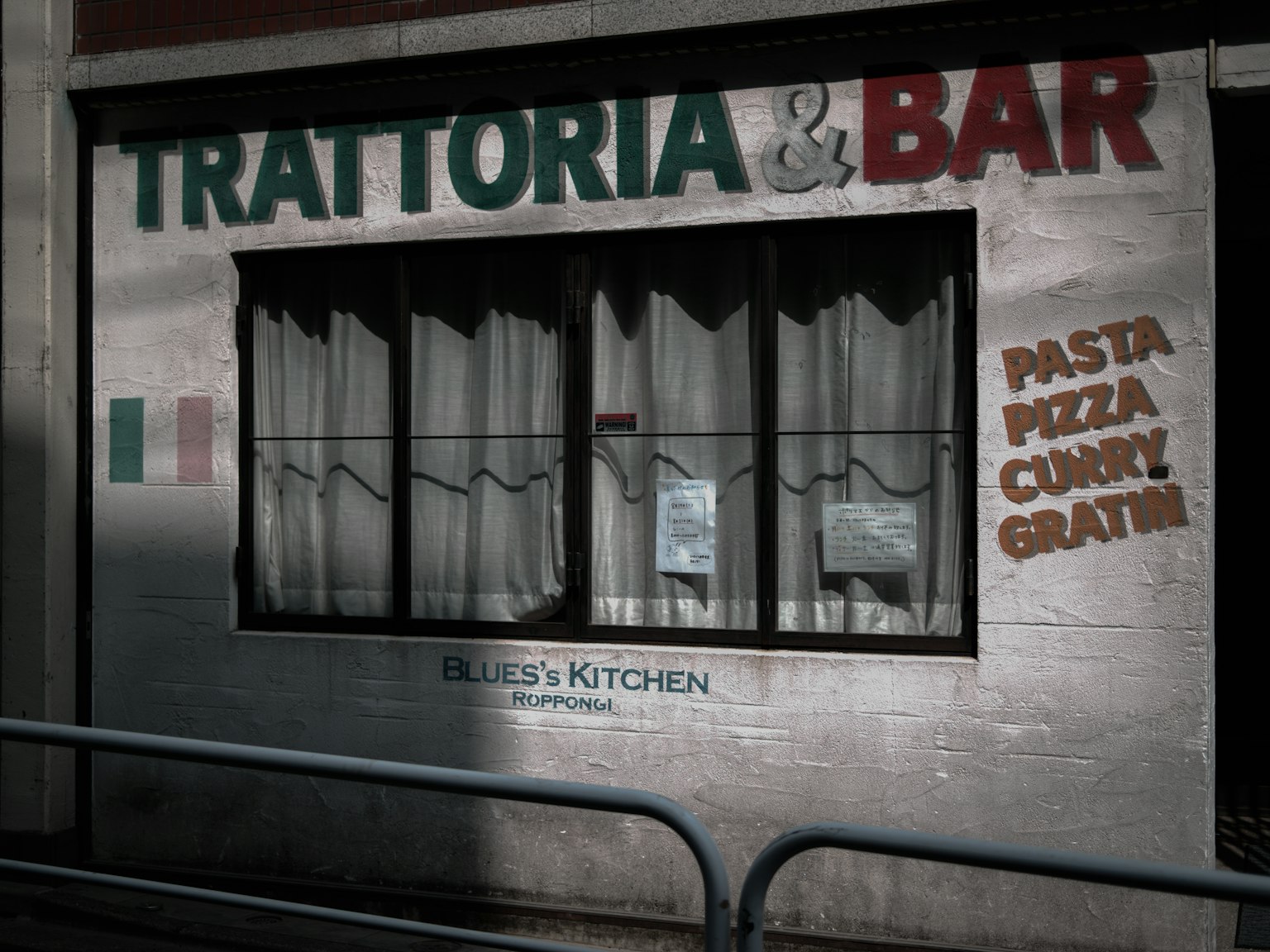 ภายนอกร้านอาหารอิตาเลียนพร้อมหน้าต่างและผ้าม่านที่มีป้าย Trattoria & Bar