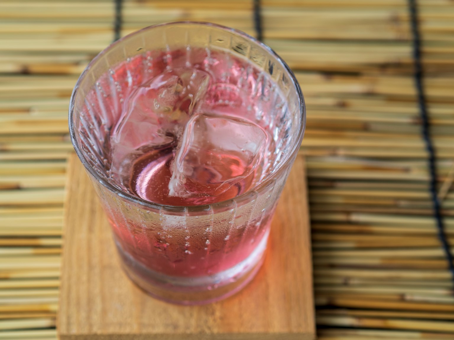 Minuman berwarna merah muda dengan es dalam gelas transparan di atas tatakan kayu