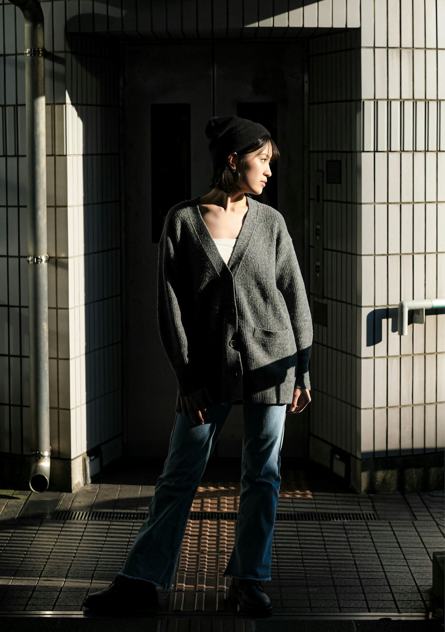 Una donna in piedi con un cardigan grigio e jeans con uno sfondo di muro piastrellato che mostra un bel contrasto di luce e ombra