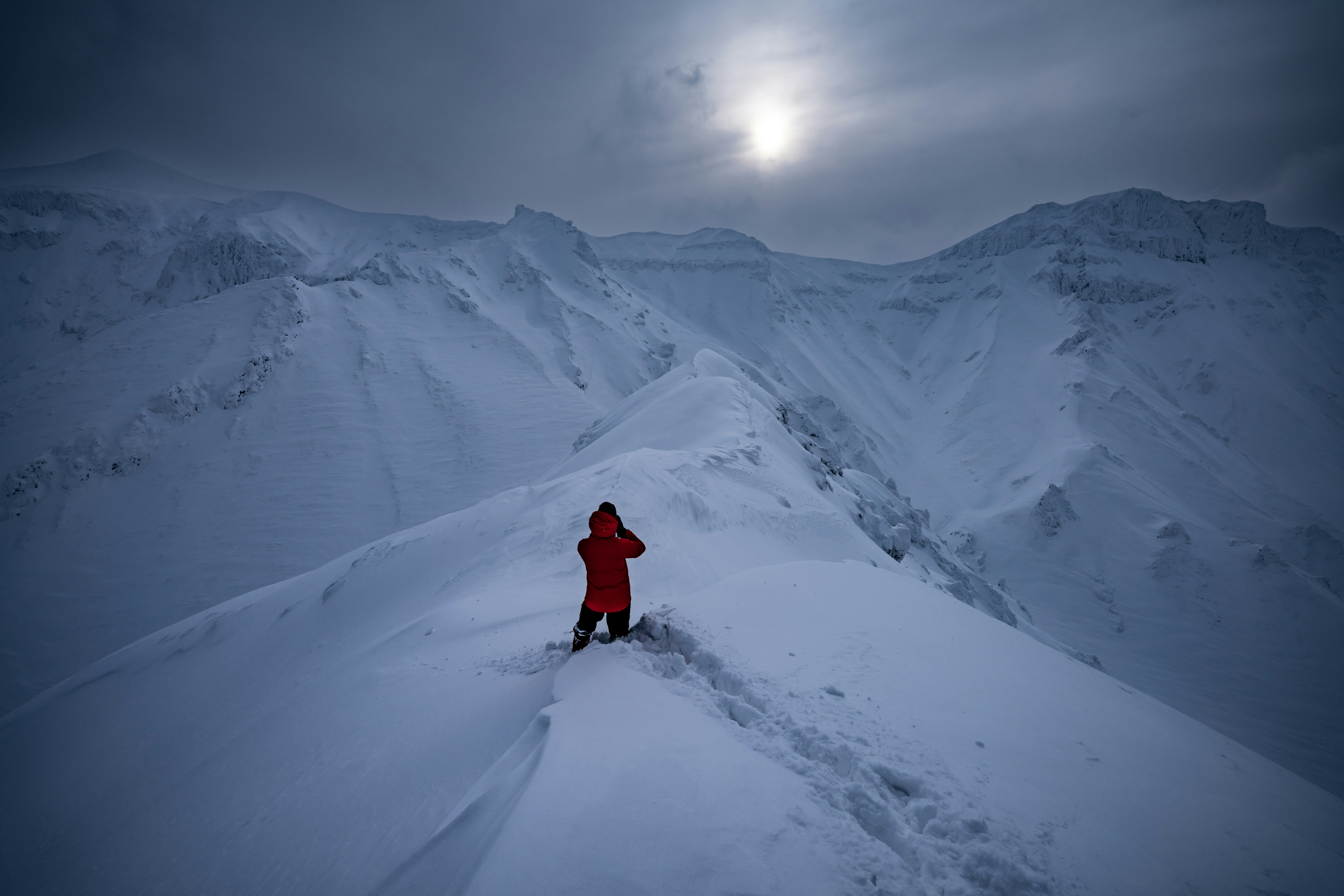 雪山の稜線を歩く赤いコートの人物と広がる山並み