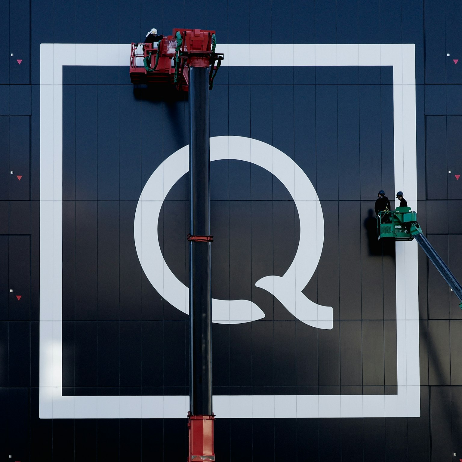 Großes weißes Q-Logo an einer schwarzen Wand mit Arbeitern auf Arbeitsbühnen