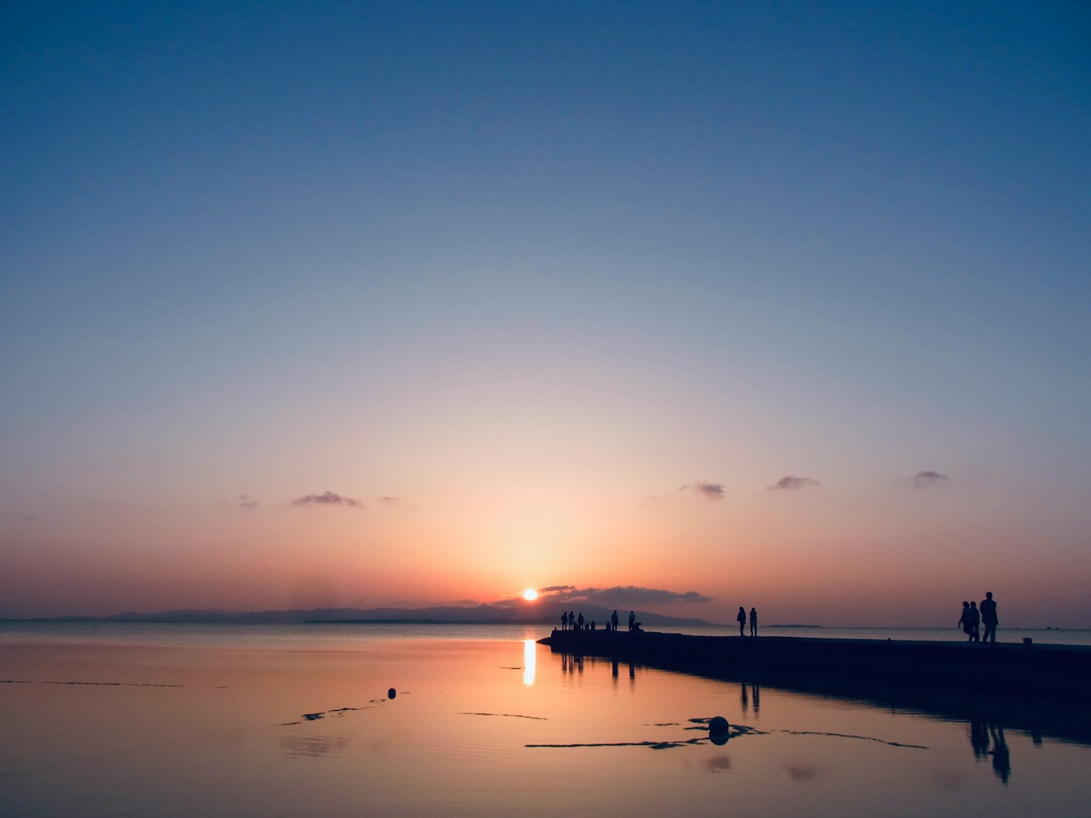 Siluetas de personas en una playa con un atardecer reflejándose en aguas tranquilas