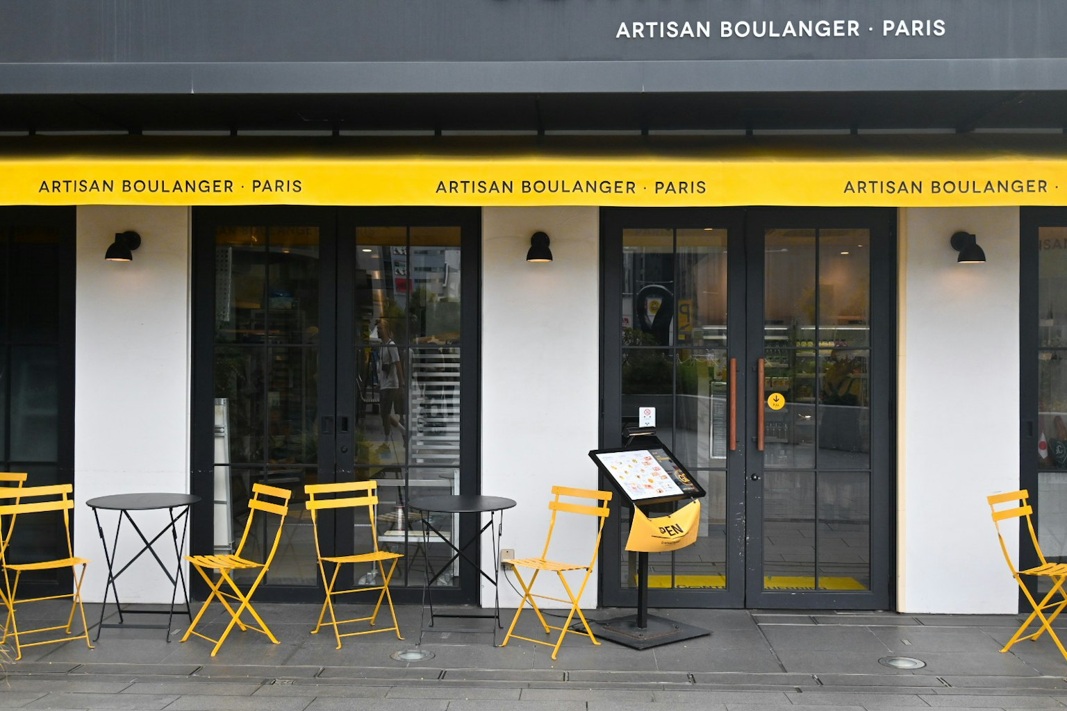 ด้านนอกของ Artisan Boulanger ในปารีสที่มีผ้าใบสีเหลืองและที่นั่งกลางแจ้ง
