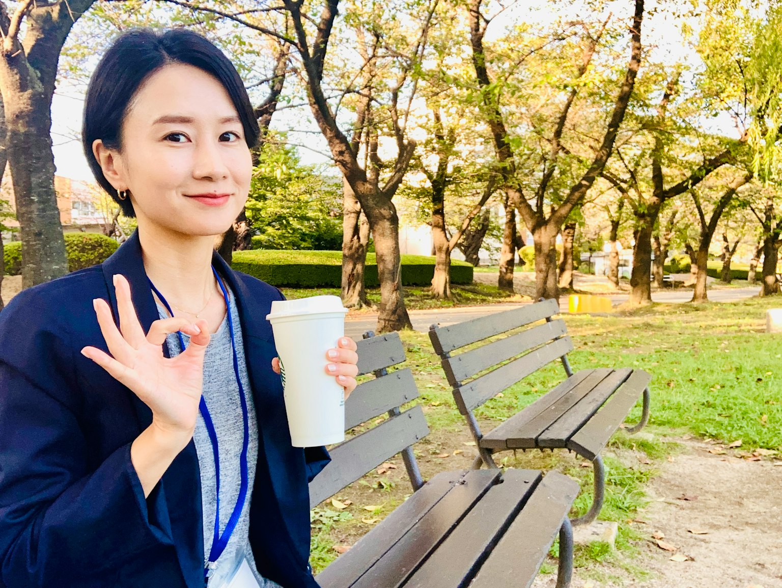 一位坐在公园长椅上的女性，手持咖啡并做出OK手势