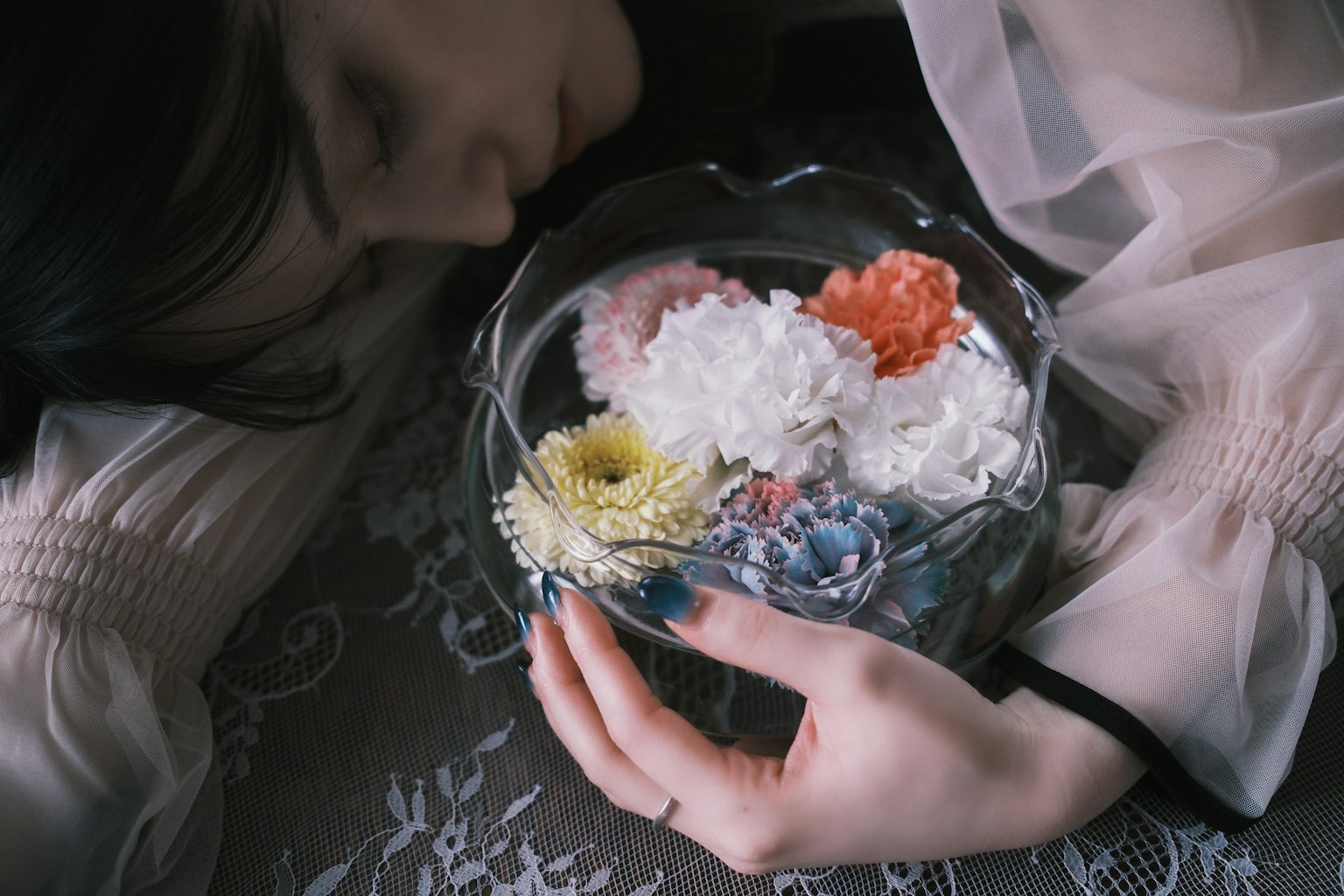 女性が花の入ったガラスの器を抱えているシーンで、柔らかい色合いの花々が特徴的