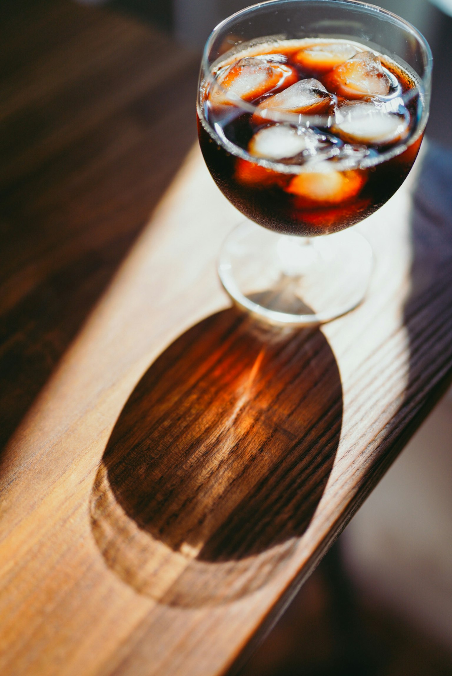 A glass of iced coffee with ice cubes casting a shadow on a wooden surface