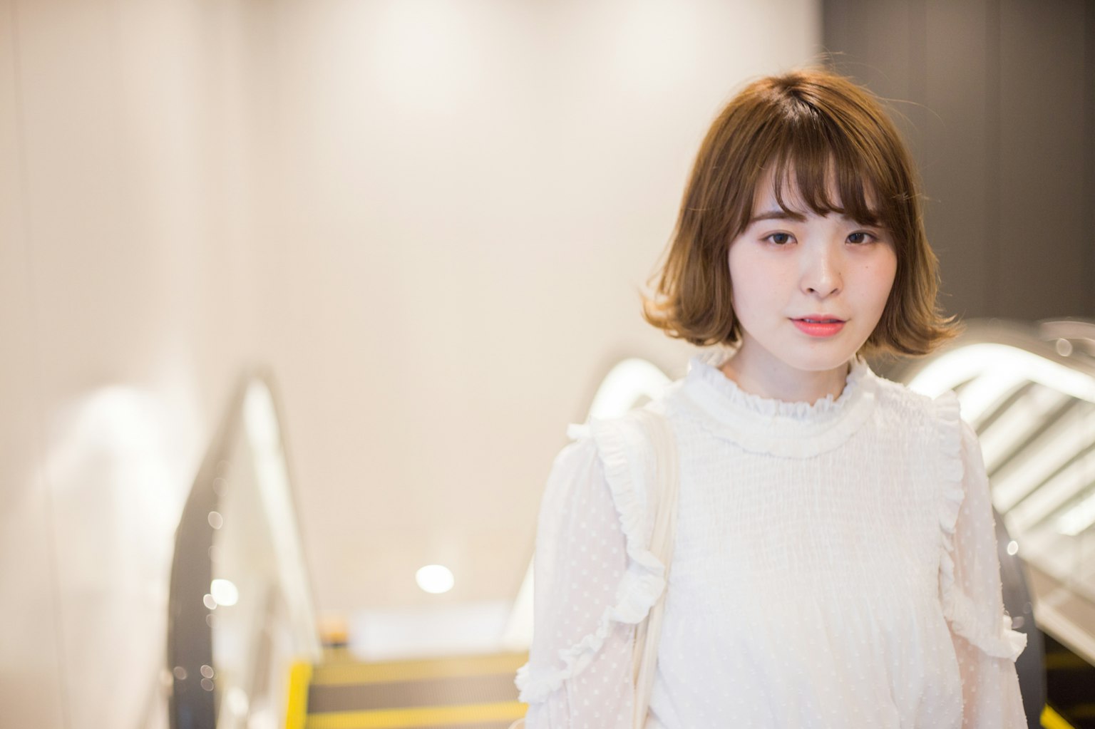 Una mujer con blusa blanca está de pie cerca de una escalera mecánica con un fondo brillante