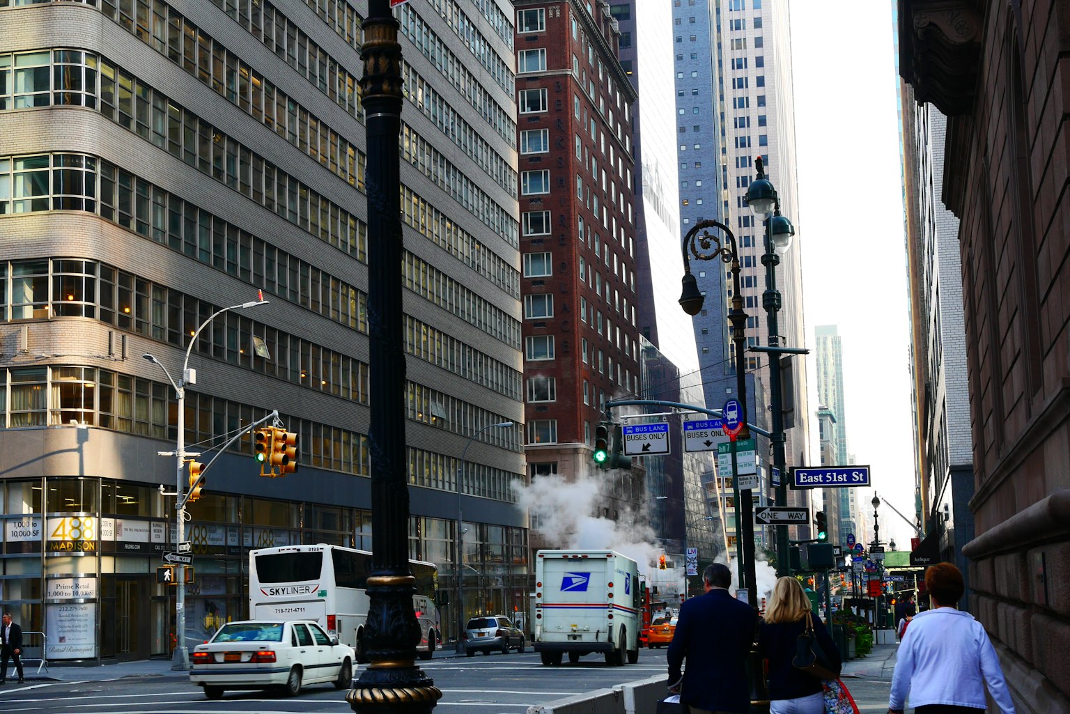 Calle de la ciudad con altos edificios y un vehículo que emite vapor junto a peatones