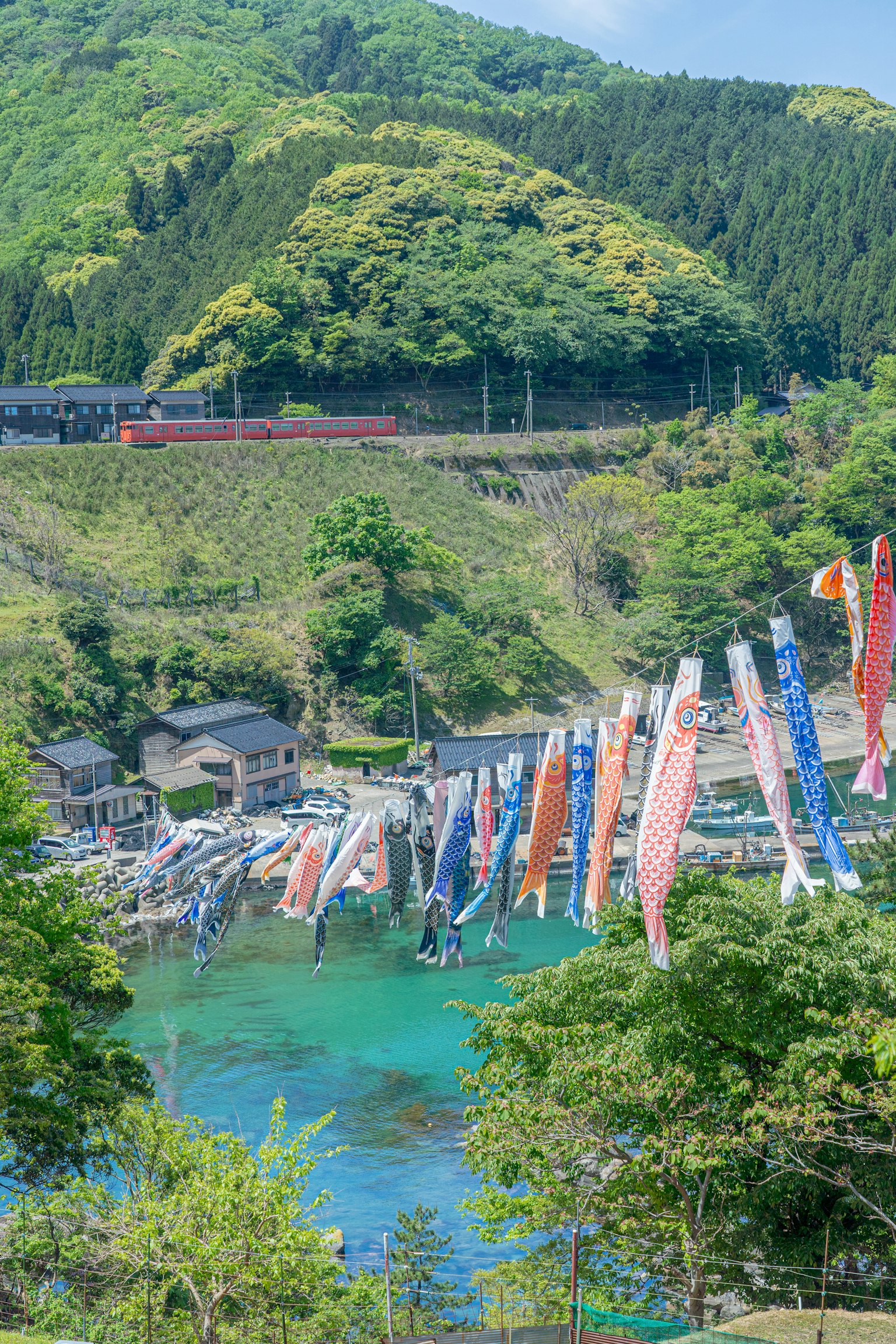 Decoraciones de koinobori coloridos junto al agua con montañas verdes y un tren rojo al fondo