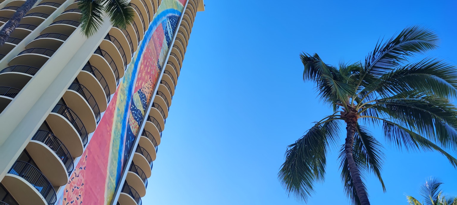 Un edificio alto con un letrero colorido contra un cielo azul y palmeras