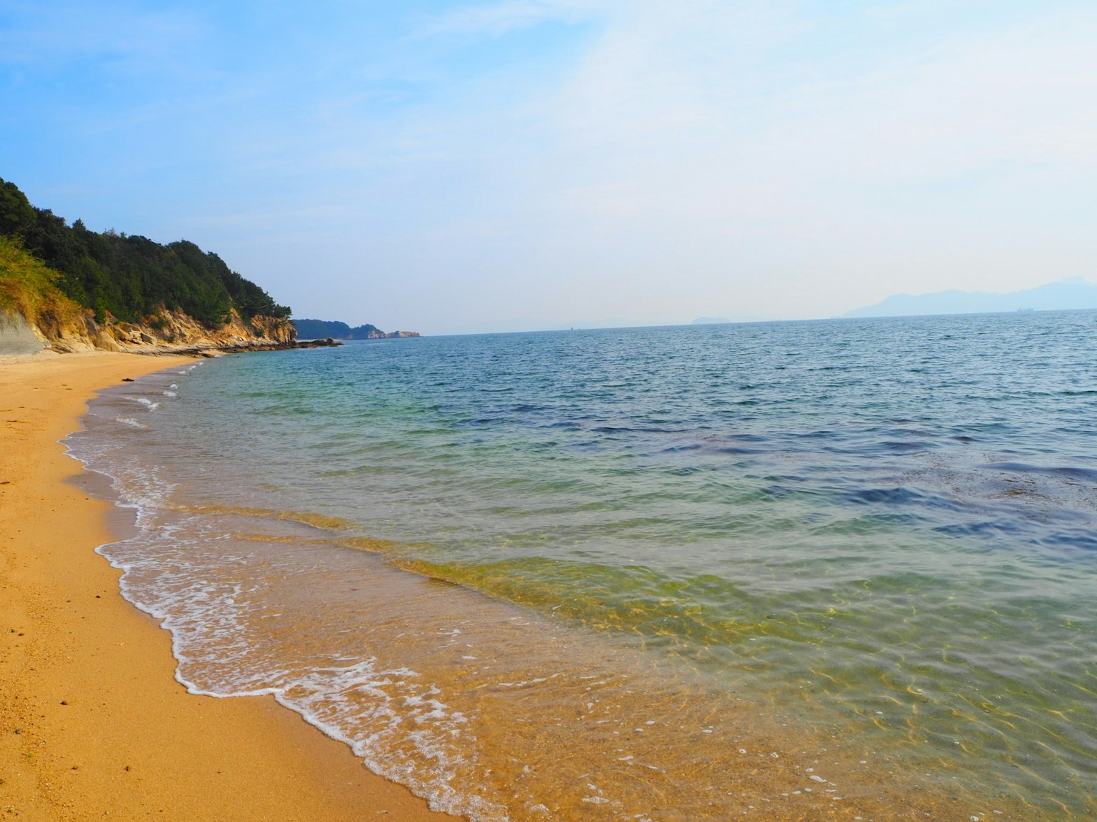 ทิวทัศน์ทะเลสงบและชายหาดทราย พร้อมเนินเขาเขียวขจีและท้องฟ้าสีฟ้า