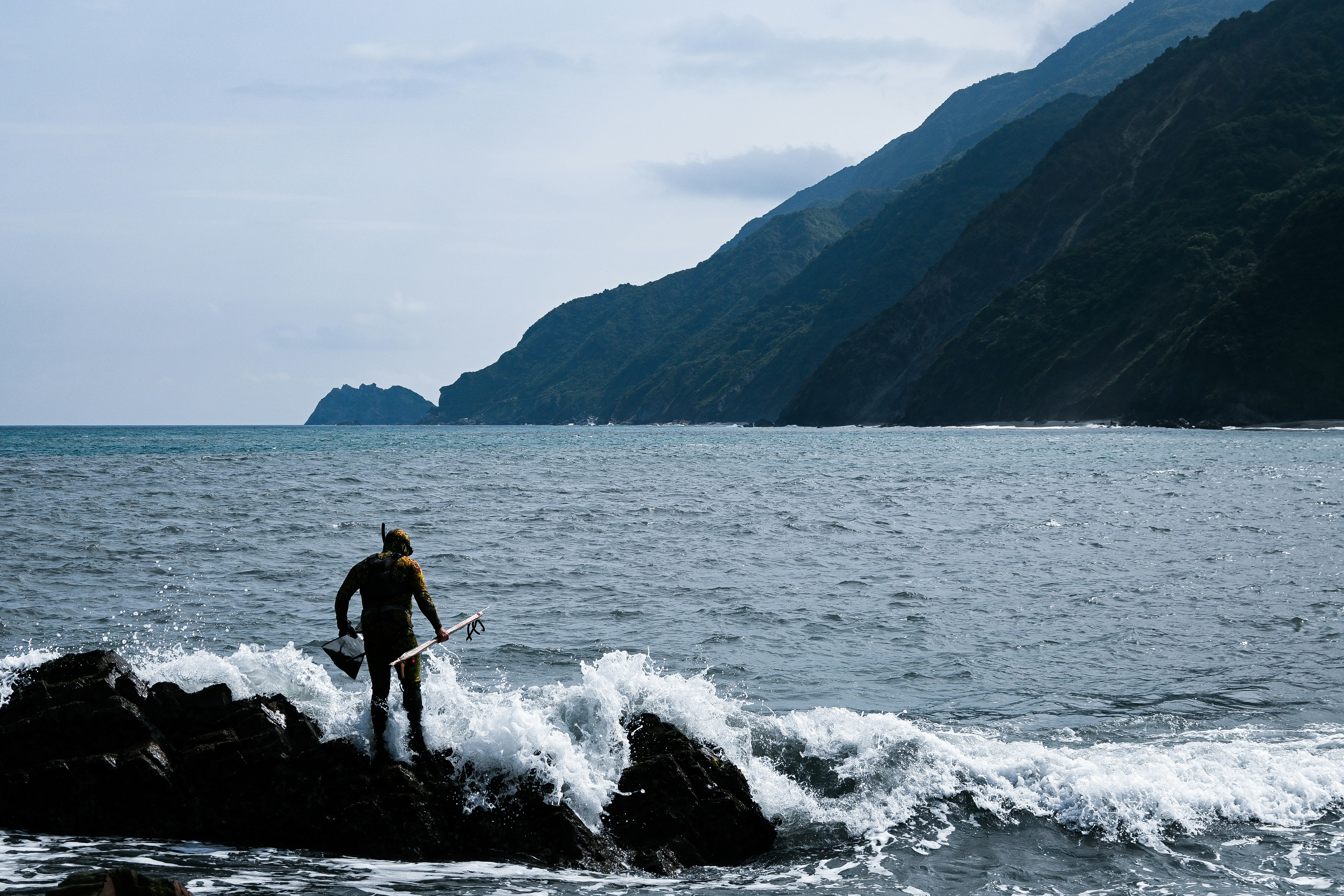 海辺の岩場で波に立つ釣り人と山並み