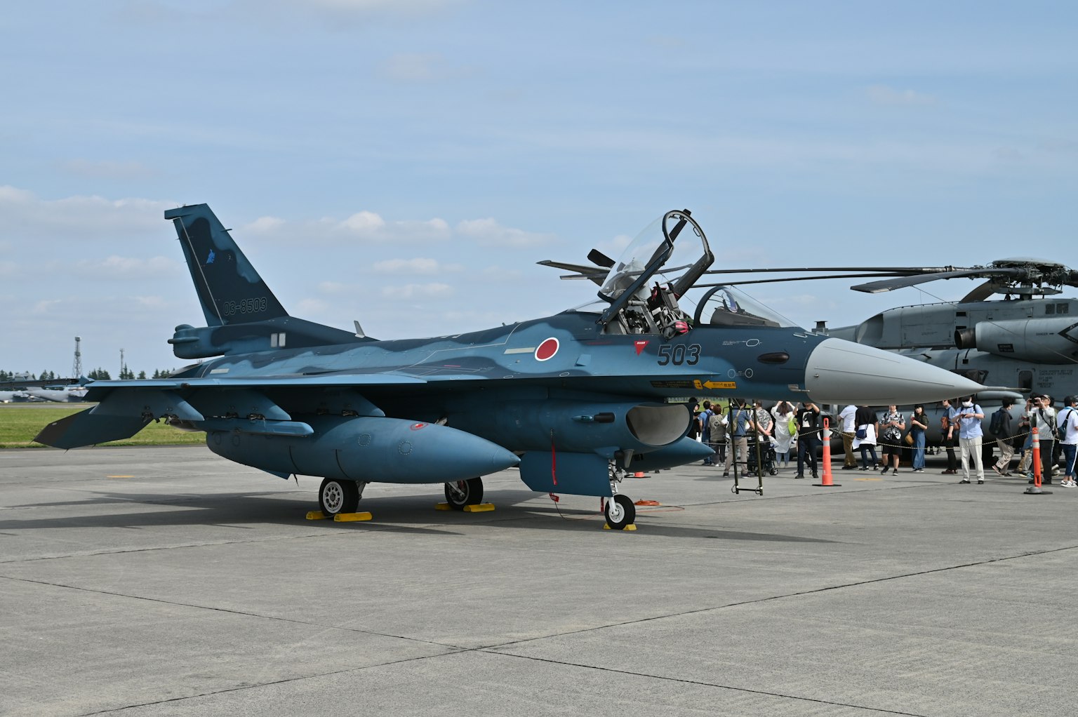 Blue fighter jet and helicopter at an airshow with spectators