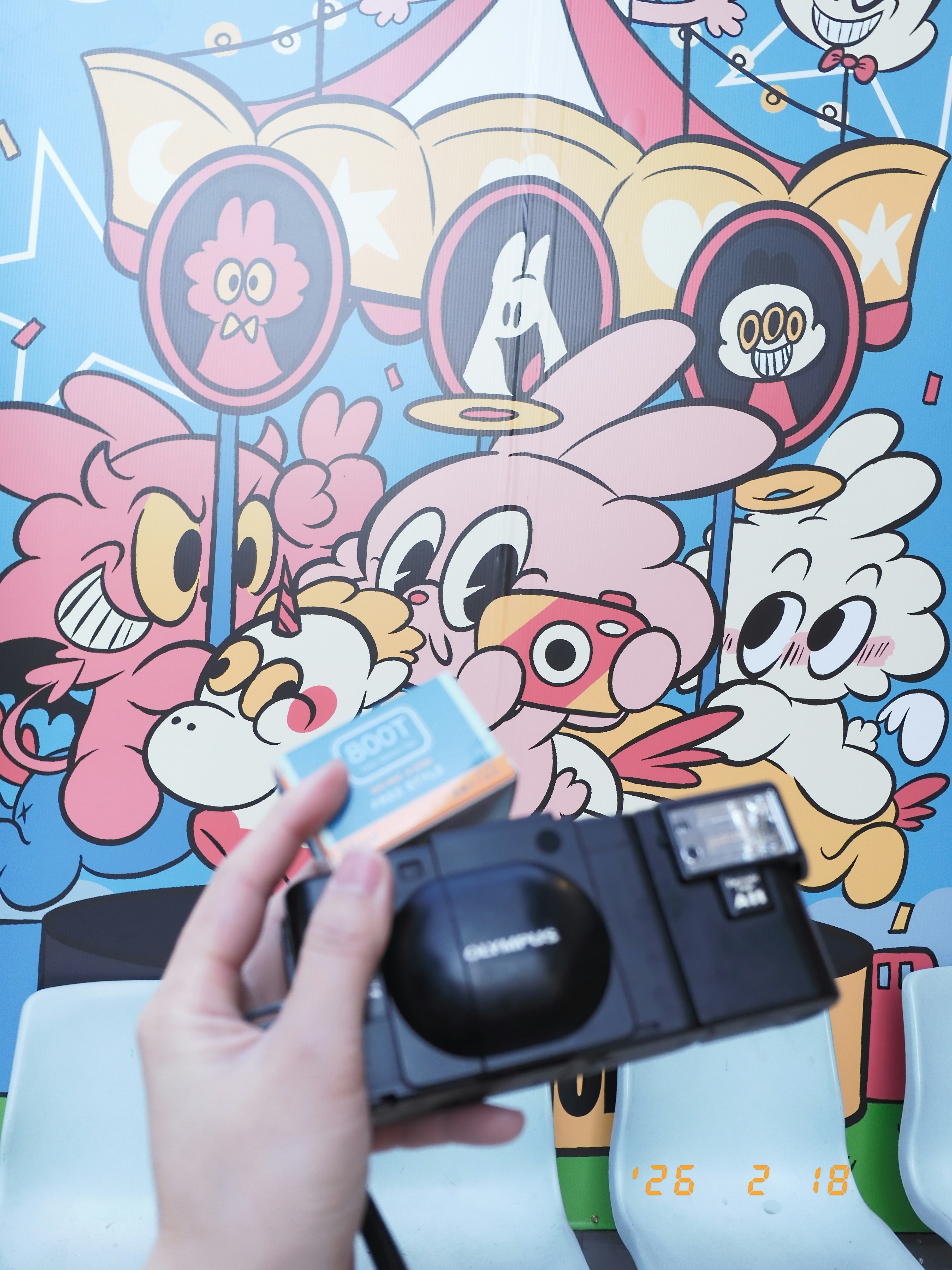 カメラとフィルムが並ぶ、カラフルなキャラクター壁面の前
