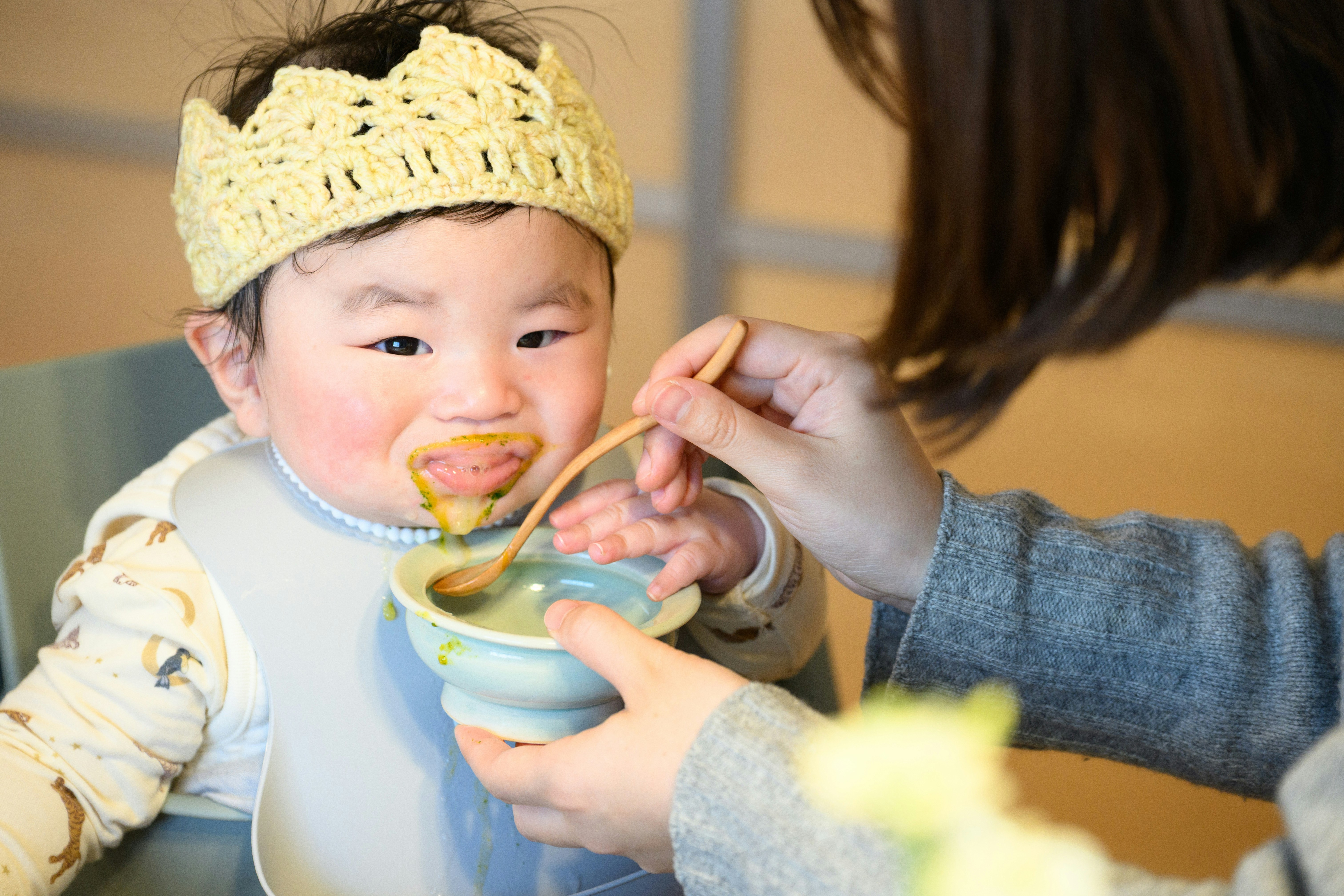 ニット帽の赤ちゃんがスプーンで食べる家族の食事風景
