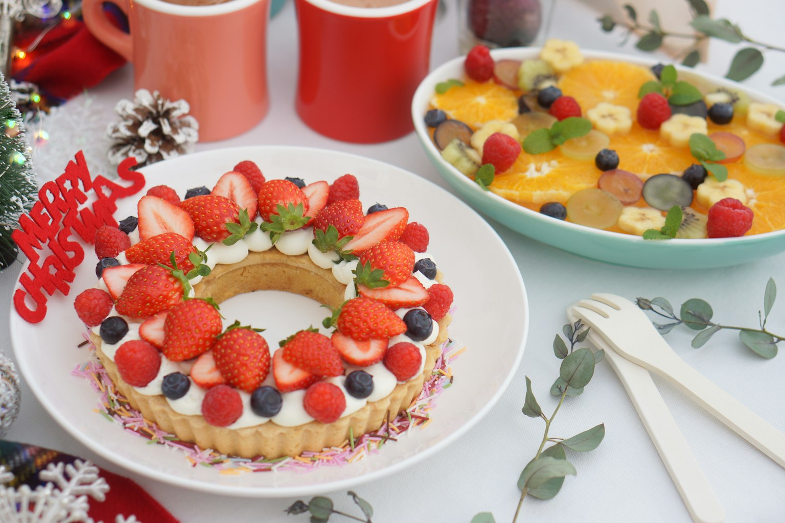 Une table festive avec un gâteau décoré de fraises et de myrtilles accompagné d'une salade de fruits colorée et des tasses rouges en arrière-plan