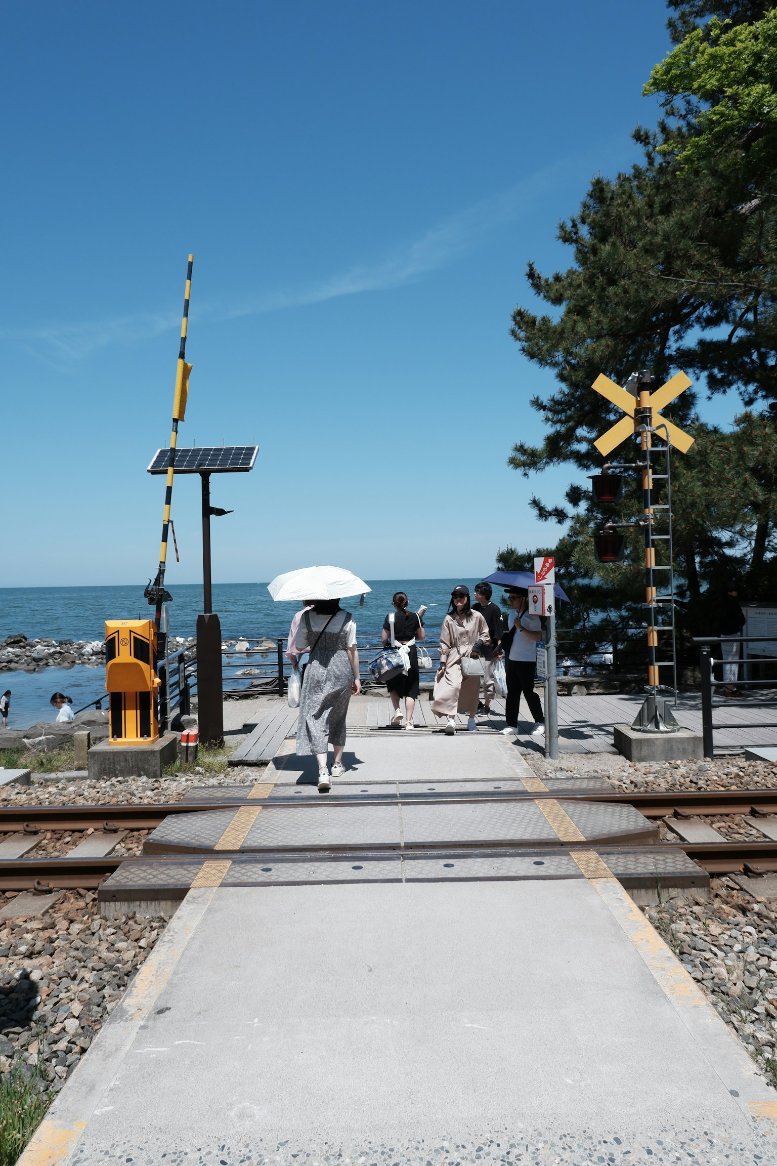 Menschen überqueren einen Bahnübergang mit blauem Himmel und Meer im Hintergrund