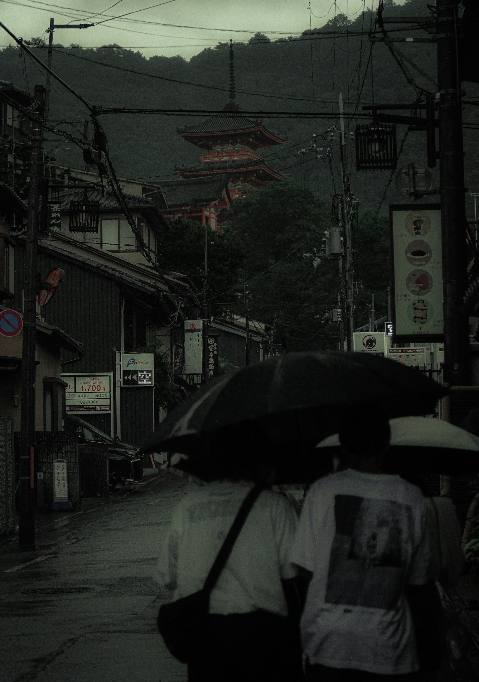 สองคนเดินถือร่มในถนนเงียบสงบในวันที่ฝนตกมีซิลลูเอตของวัดอยู่เบื้องหลัง
