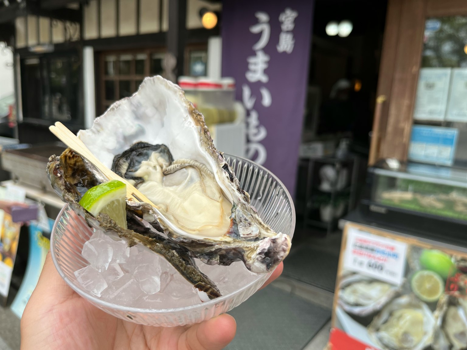 A hand holding a fresh oyster with a slice of lime in a clear dish in front of a Japanese restaurant sign