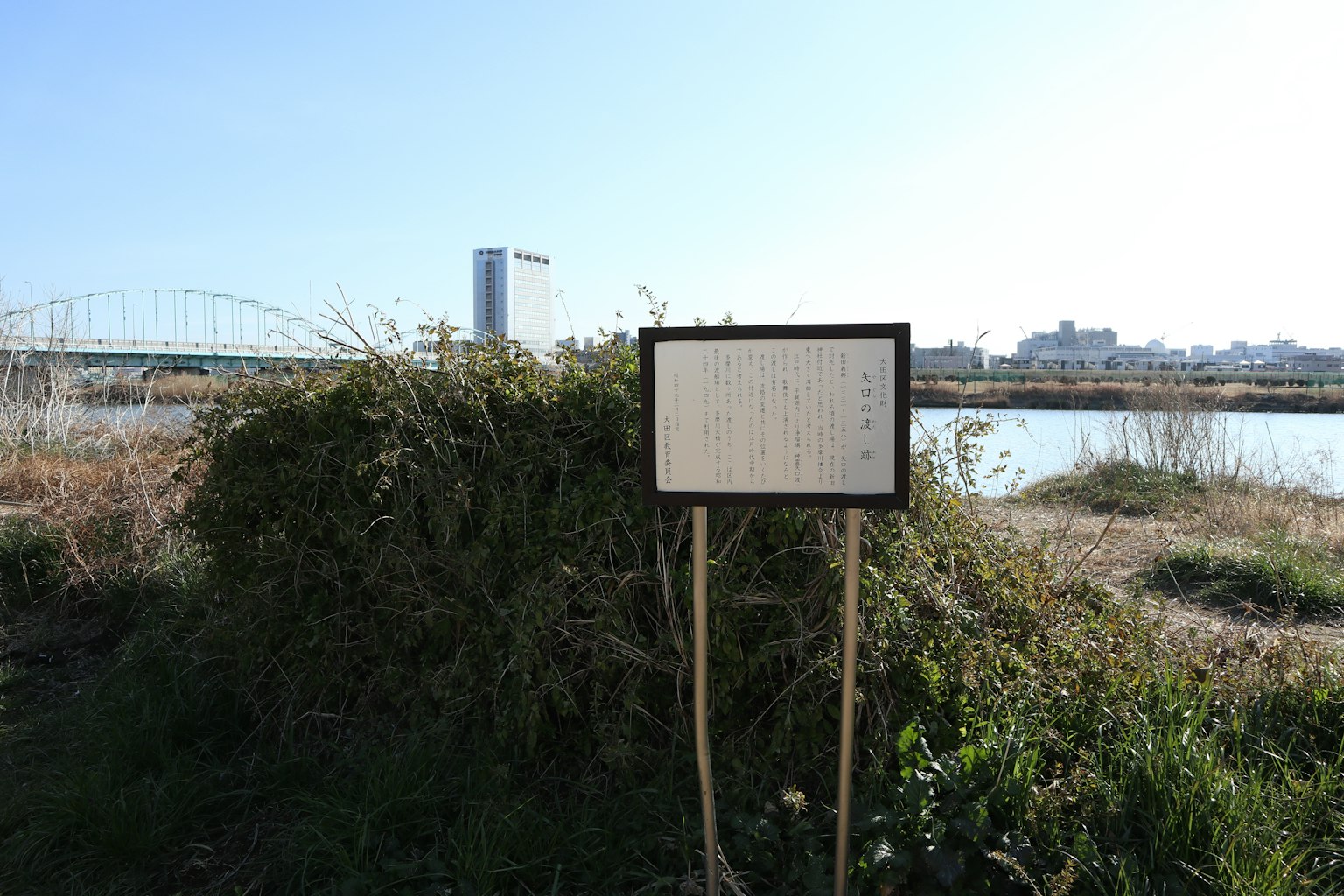 Un panneau se tenant dans une zone herbeuse au bord de la rivière avec un bâtiment au loin