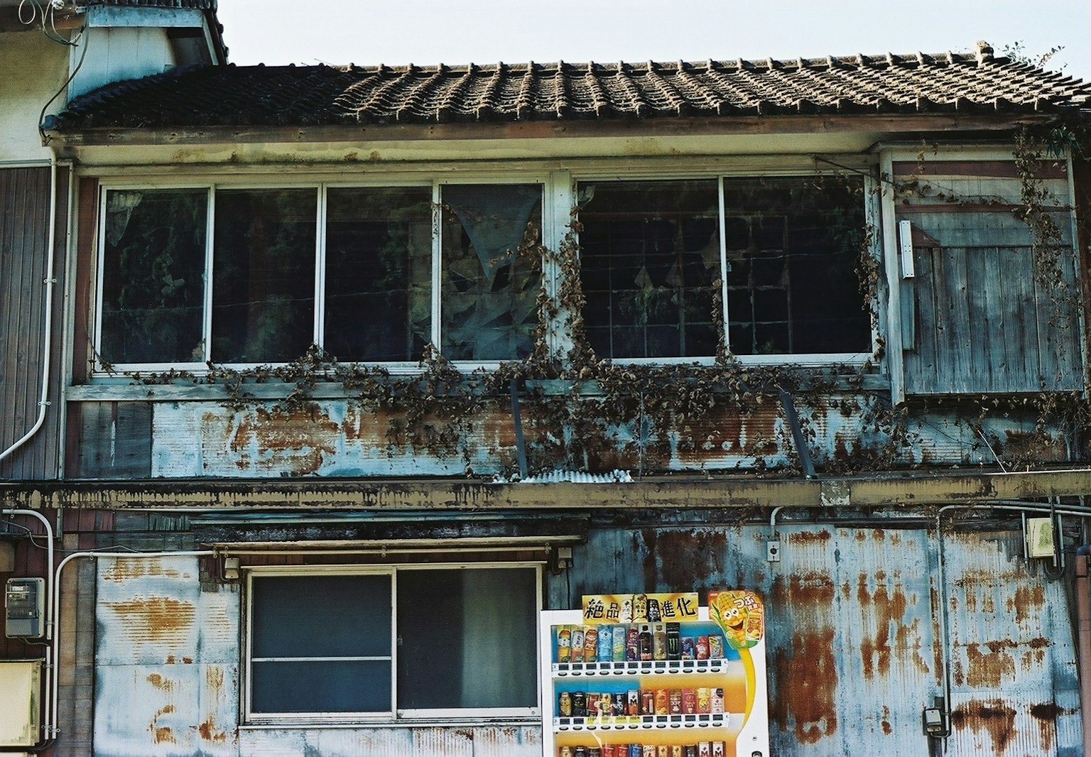 Exterior of an old building with rusted walls and windows covered in ivy and a vending machine
