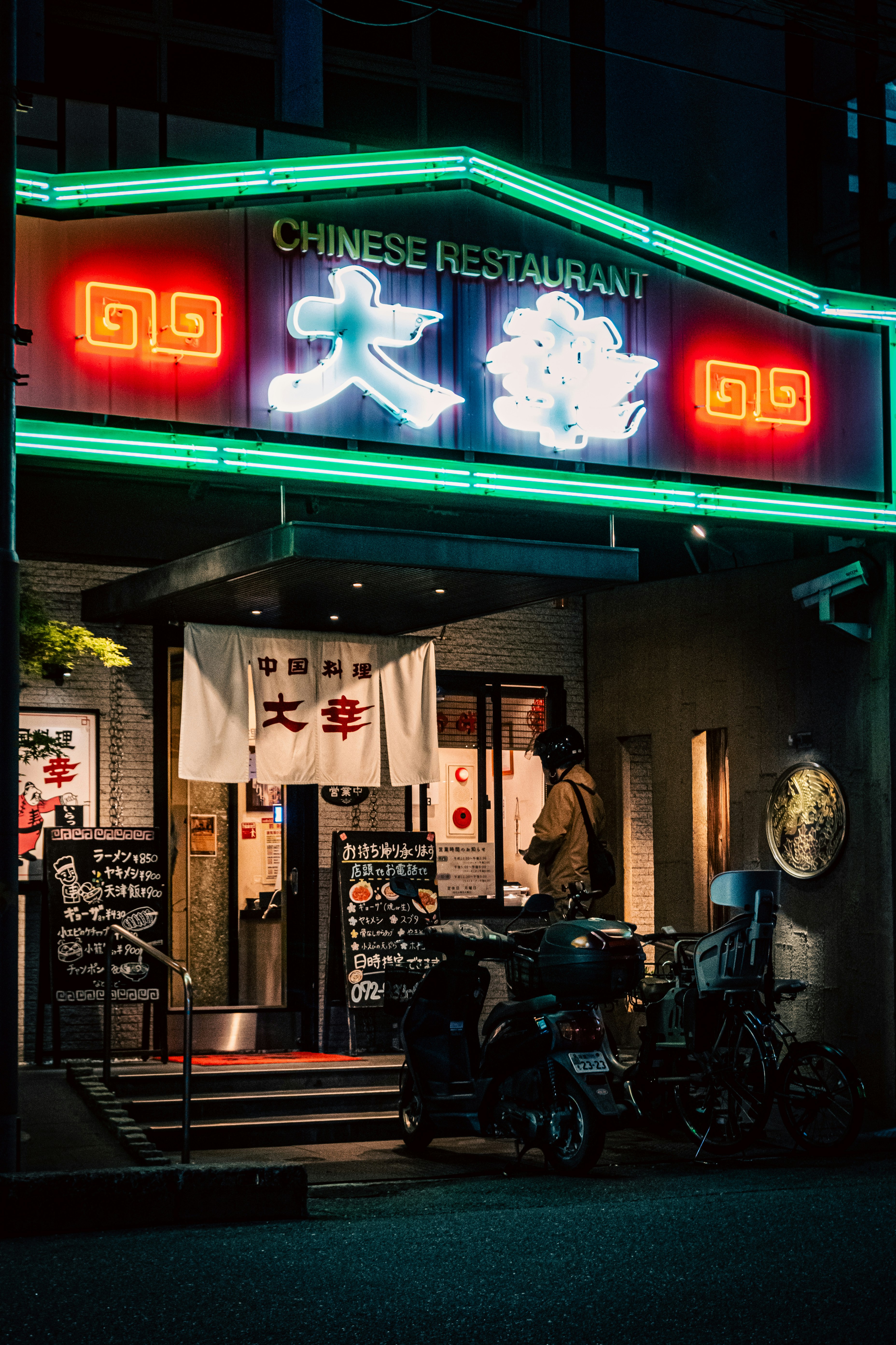 ネオンの看板が目立つ中華レストランの入口とバイクが並ぶ路地