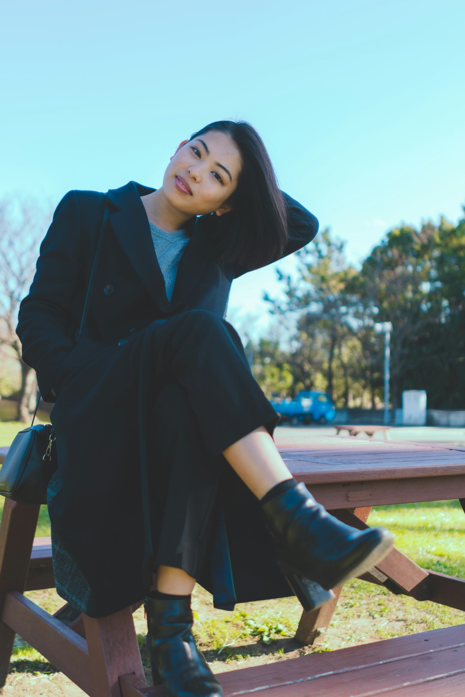 Ritratto di una donna seduta su una panchina del parco con un cappotto nero e stivali sorridendo mentre sistema i capelli