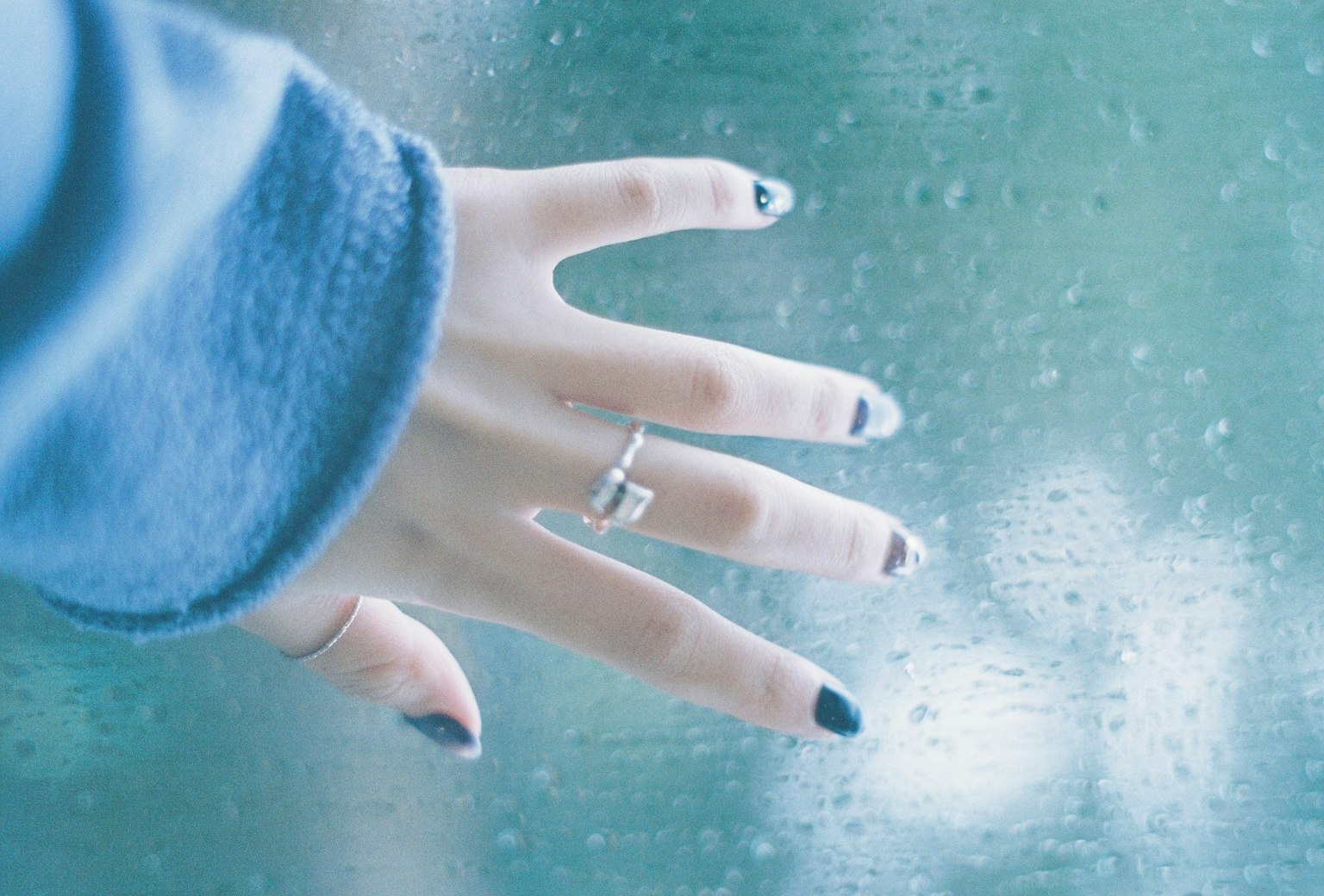 Nahaufnahme einer Hand, die ein regennasses Fenster berührt, trägt einen blauen Pullover