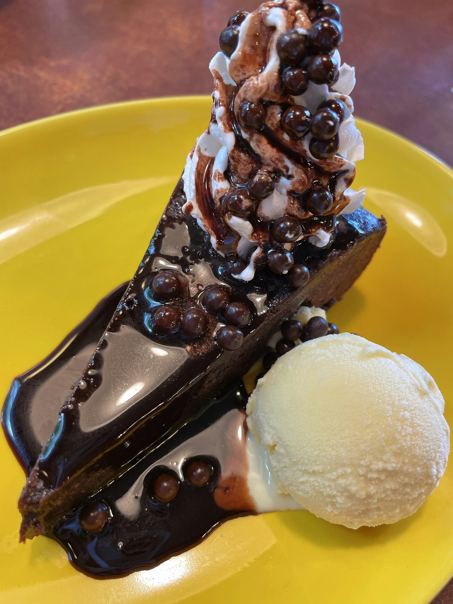 Dessert sur une assiette jaune avec un gâteau au chocolat surmonté de crème fouettée et de sauce au chocolat accompagné d'une boule de glace à la vanille