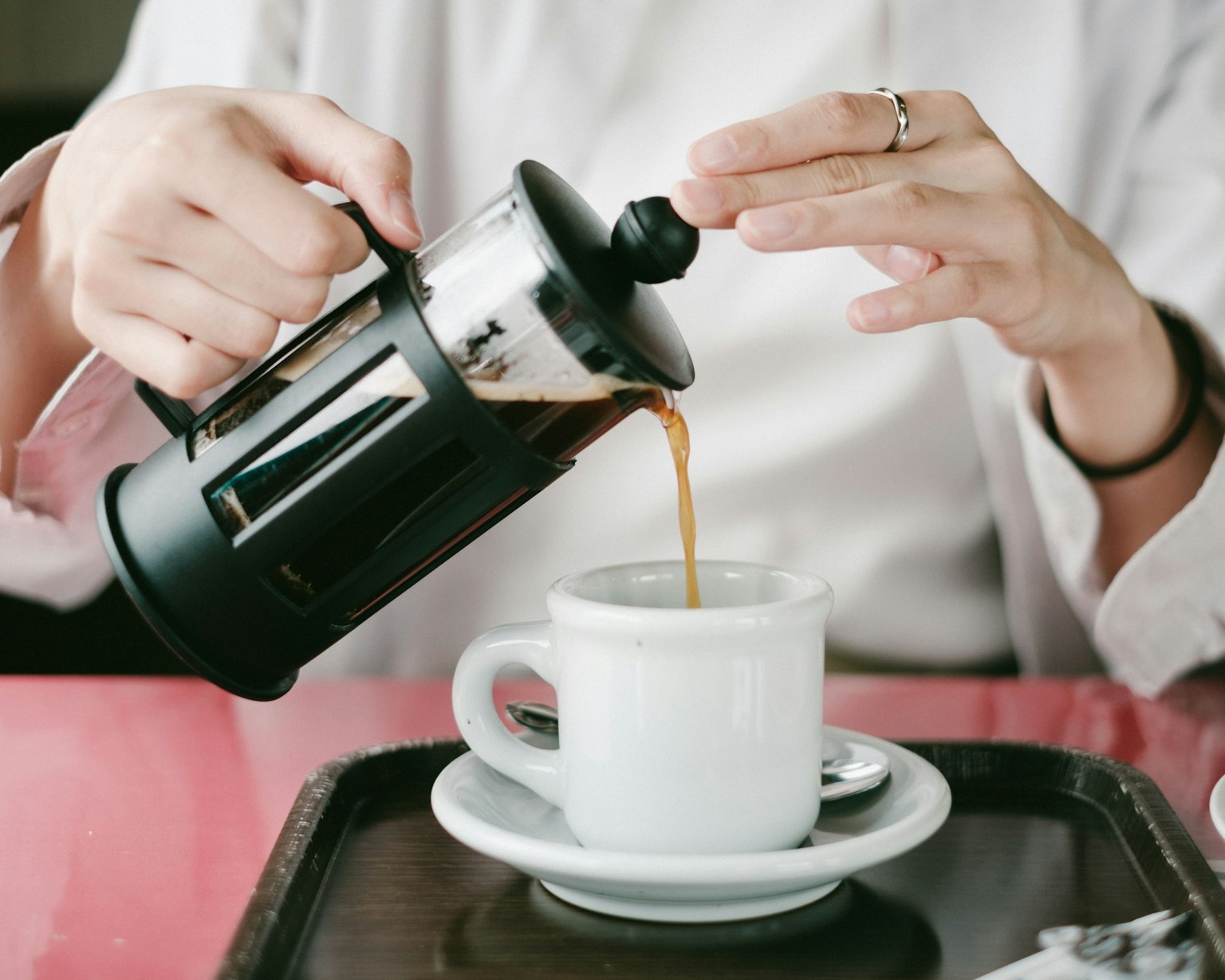 A close-up of hands pouring coffee from a French press into a white cup